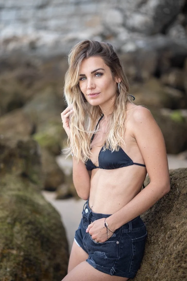 A woman in a black bikini top and black denim shorts standing outdoors next to rocks with a blurred natural background.
