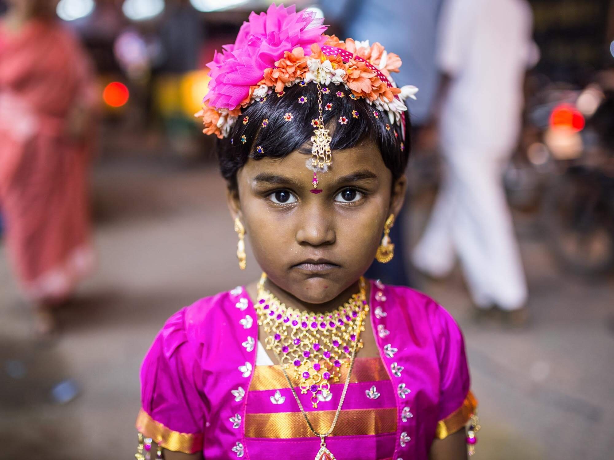 A young girl in traditional Indian attire, adorned with jewelry, wearing a colorful dress and a floral headpiece, looking directly at the camera with a serious expression. Blurred background with people and street scene.