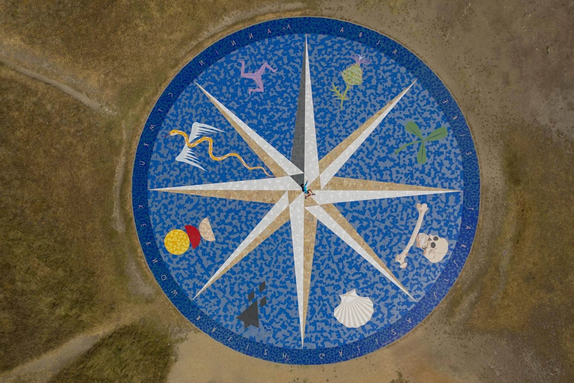 A circular outdoor mosaic artwork resembling a clock face, decorated with colorful illustrations of sea creatures, a skeleton, a seashell, a plant, and geometric shapes, on a blue background with an alphabet ring around the edge.