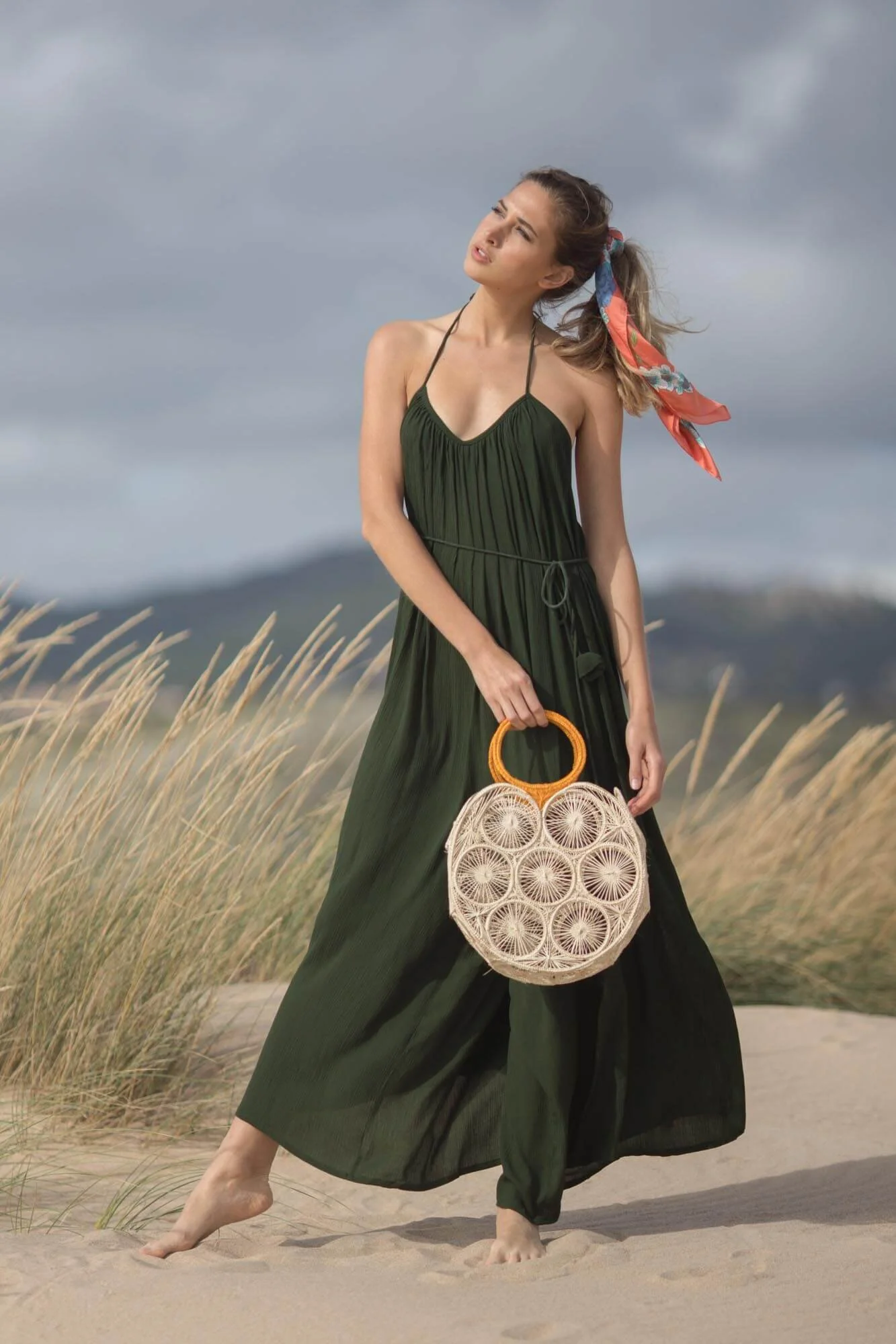 A woman in a green dress holding a round woven bag, standing barefoot on a sandy beach with tall grasses and an overcast sky.