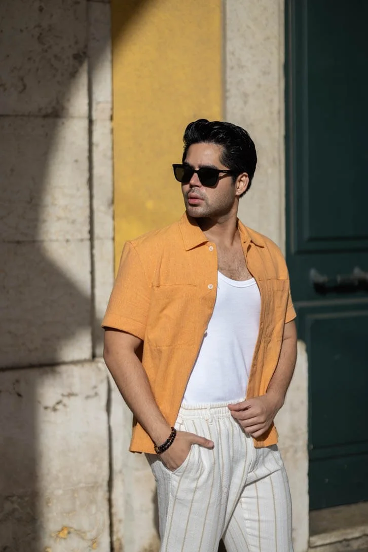 A young man with dark hair wearing black sunglasses, an orange short-sleeve button-up shirt over a white tank top, and white striped pants stands outdoors against a wall with a green door, looking to the side.