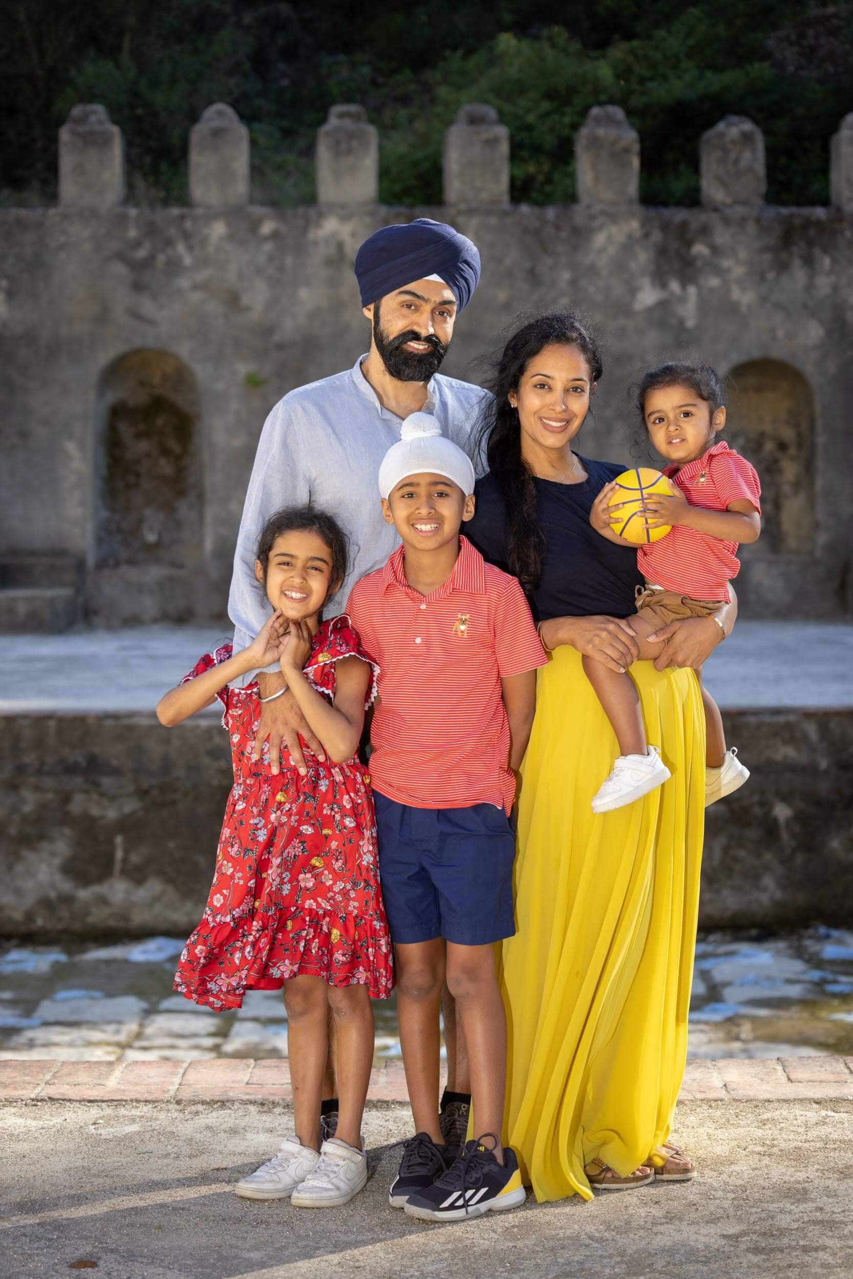 A family of five posing outdoors near a historic stone wall, with trees in the background. The father is wearing a blue turban and light blue shirt, the mother in a black top and yellow skirt holding a young girl in red striped shirt and beige shorts