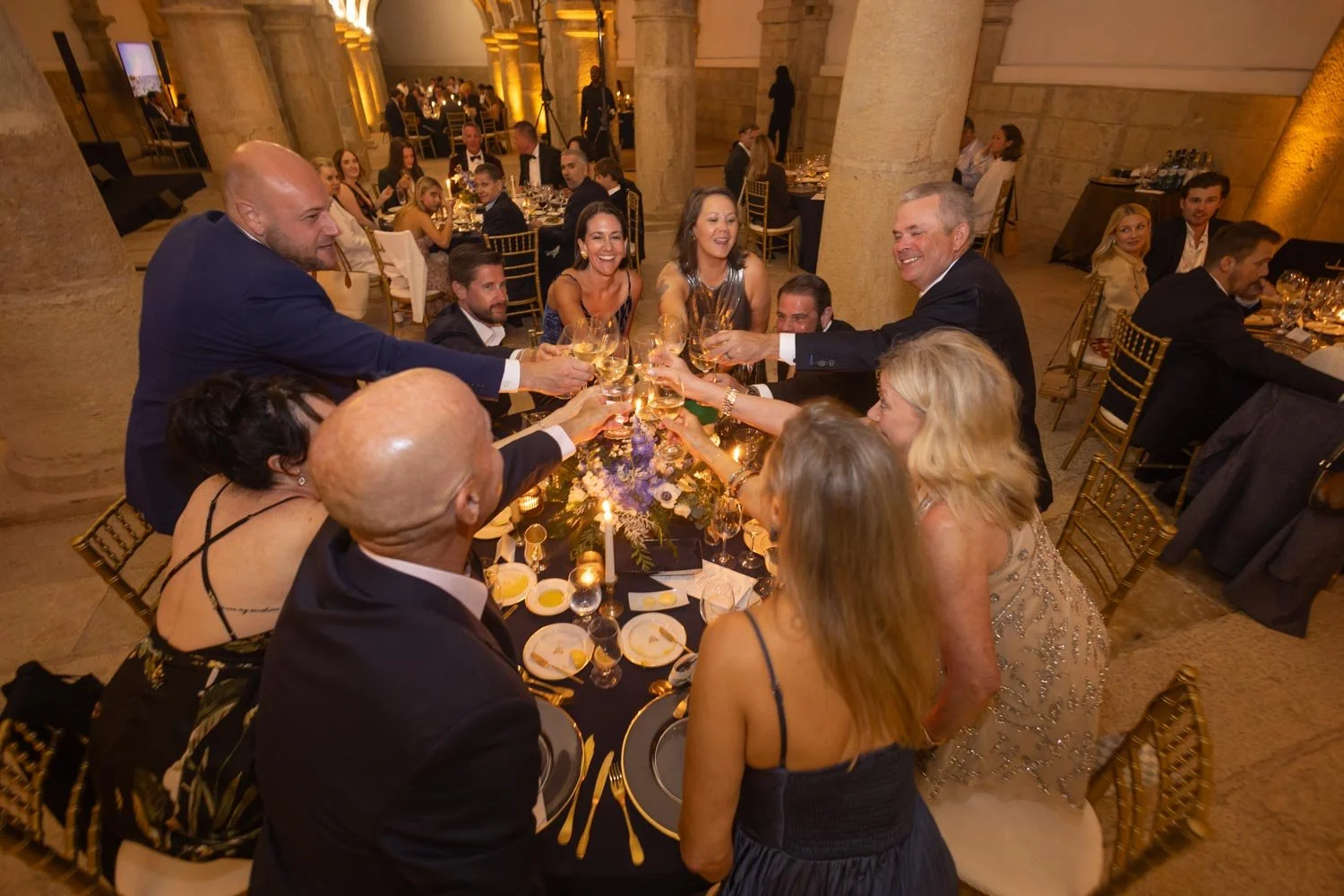 A group of people dressed in formal attire raising glasses in a toast at a celebration dinner in an elegant, warmly lit banquet hall with stone pillars and tables decorated with flowers and candles.