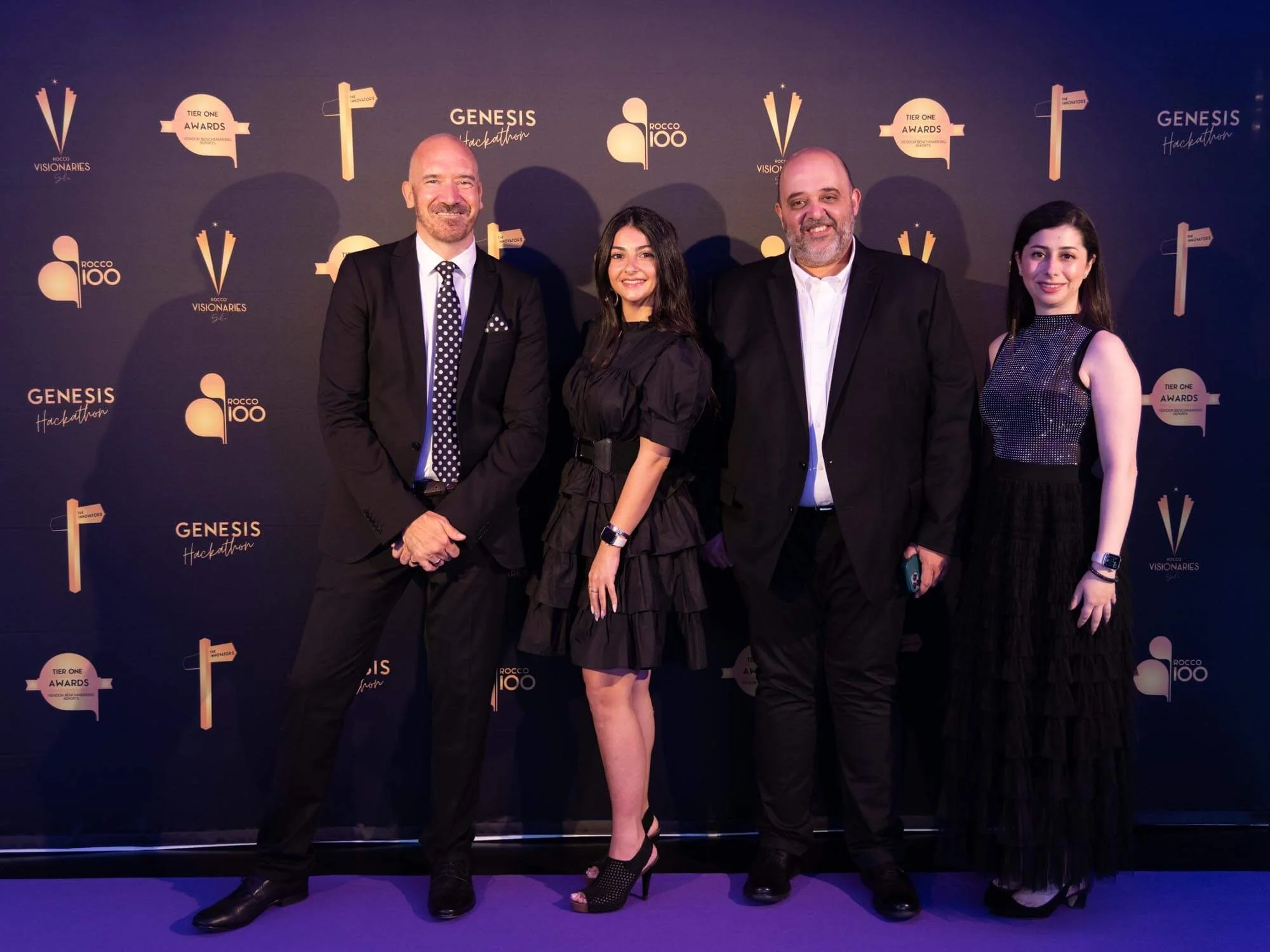 Group of four people standing on purple carpet in front of a dark backdrop with logos and text, dressed in formal attire at an event.