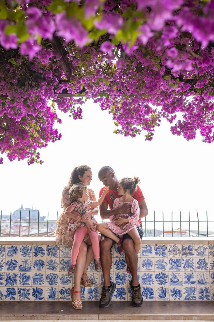 Four children sitting on a blue and white tiled wall beneath pink flowering trees, with a city skyline in the background.