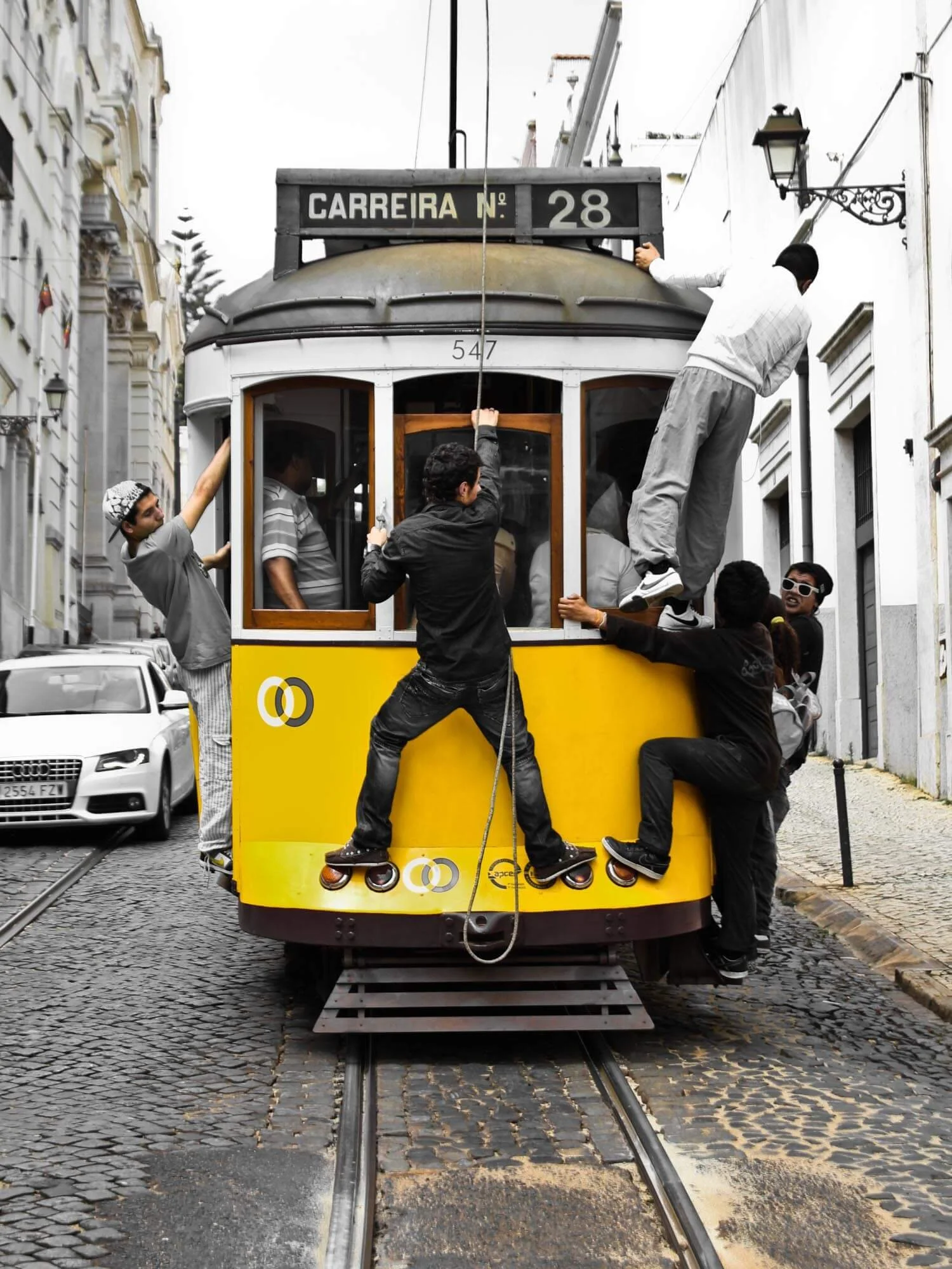 People climbing and hanging onto the front of a yellow tram on narrow city street
