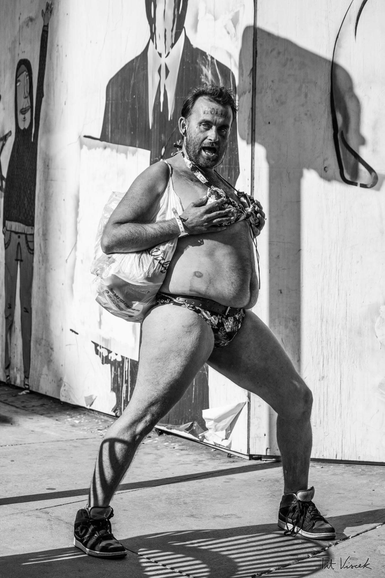 A man standing outdoors against a wall with graffiti, wearing minimal clothing including shorts, sneakers, and a chest harness, with a plastic bag hanging from his shoulder, making a provocative expression.