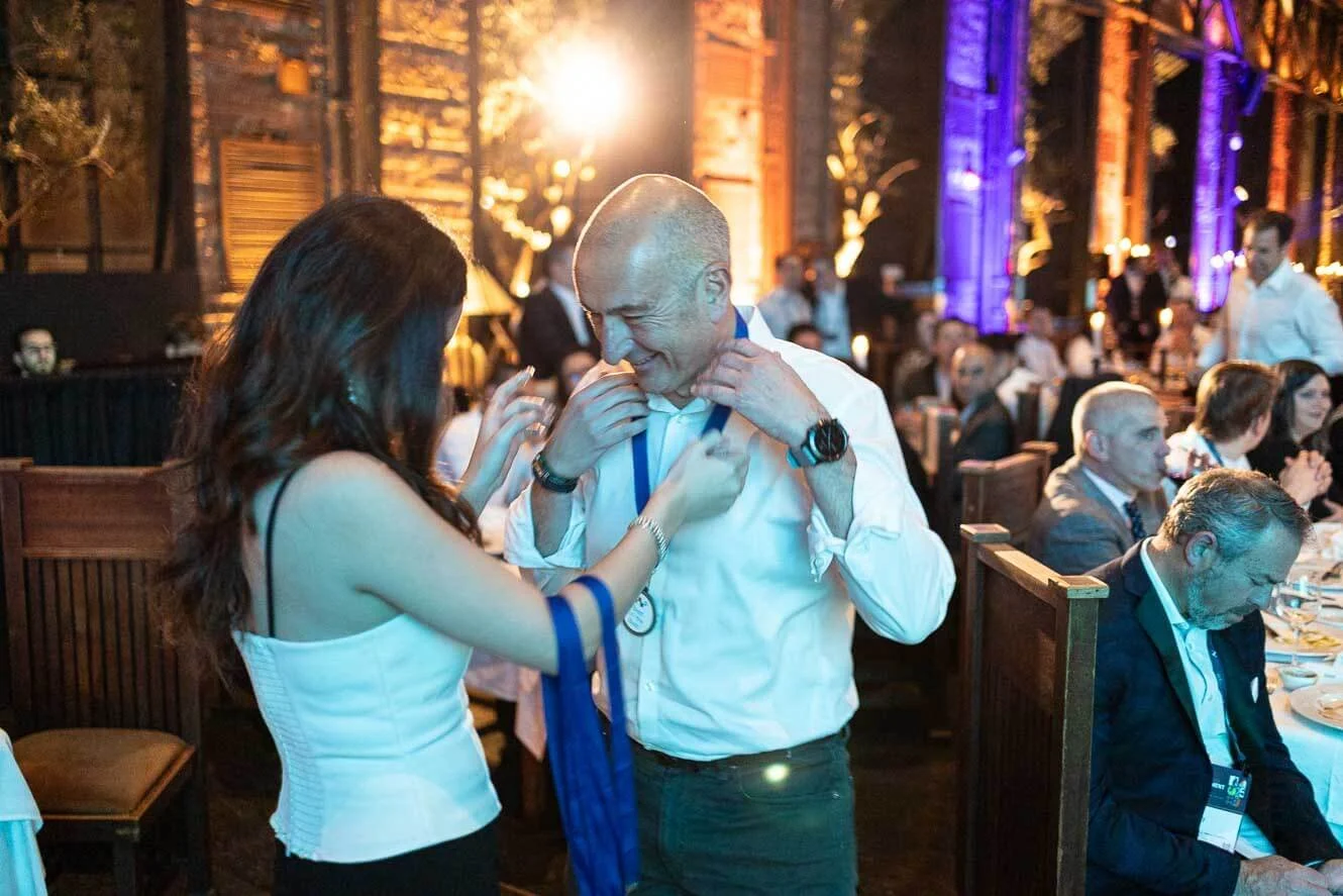 A woman is helping a man put on a medal or lanyard in a warmly lit, decorated event hall filled with seated guests.