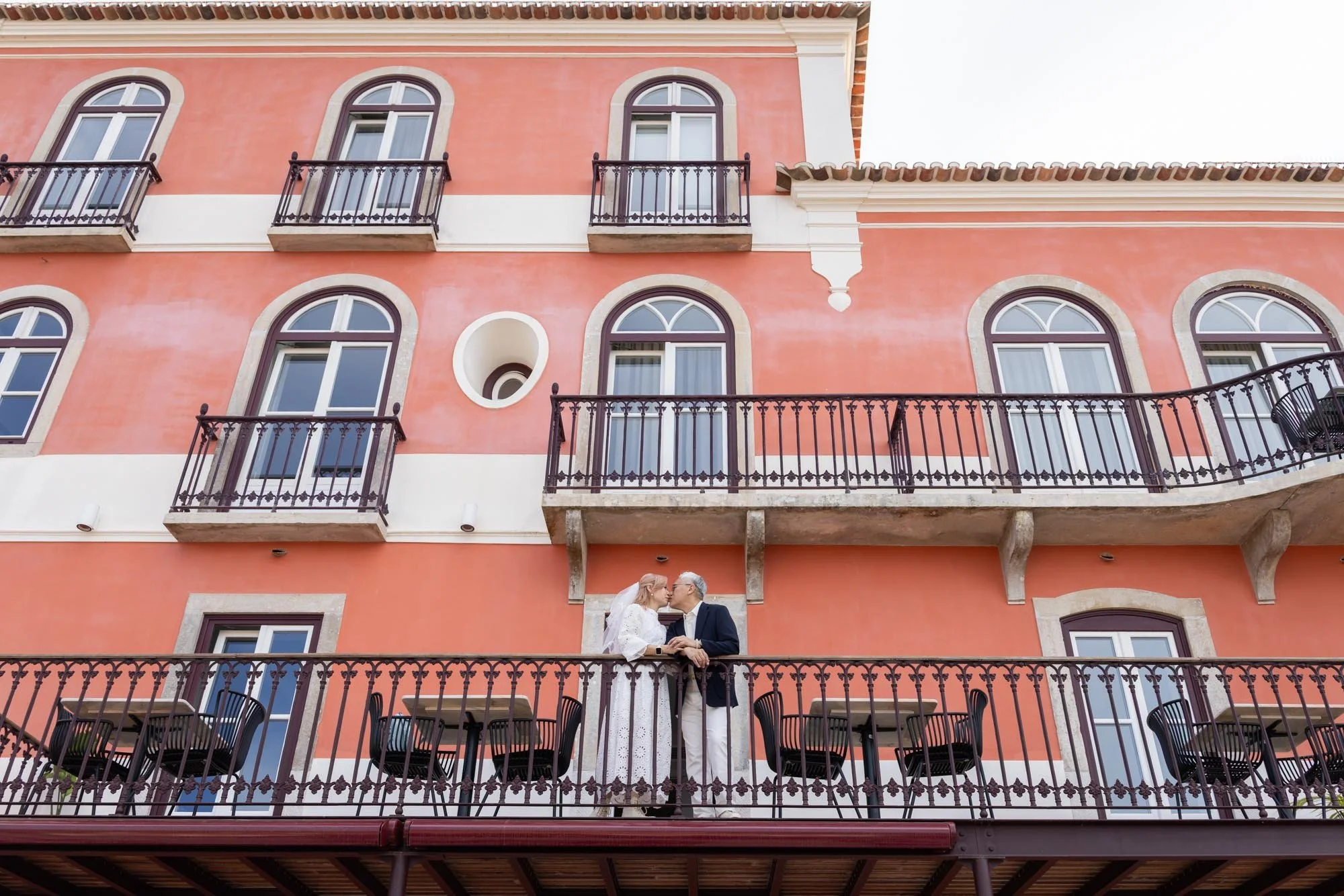 A couple standing on a balcony of a pink building, sharing an intimate moment. The building has multiple arched windows and balconies with decorative iron railings.