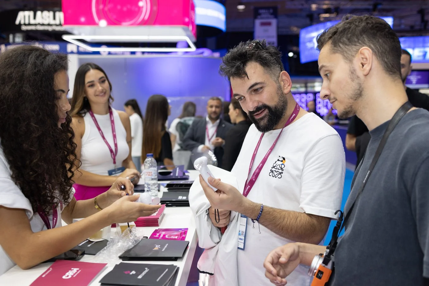 People at a technology trade show booth, examining a small white gadget, with brochures, water bottles, and lanyards visible.