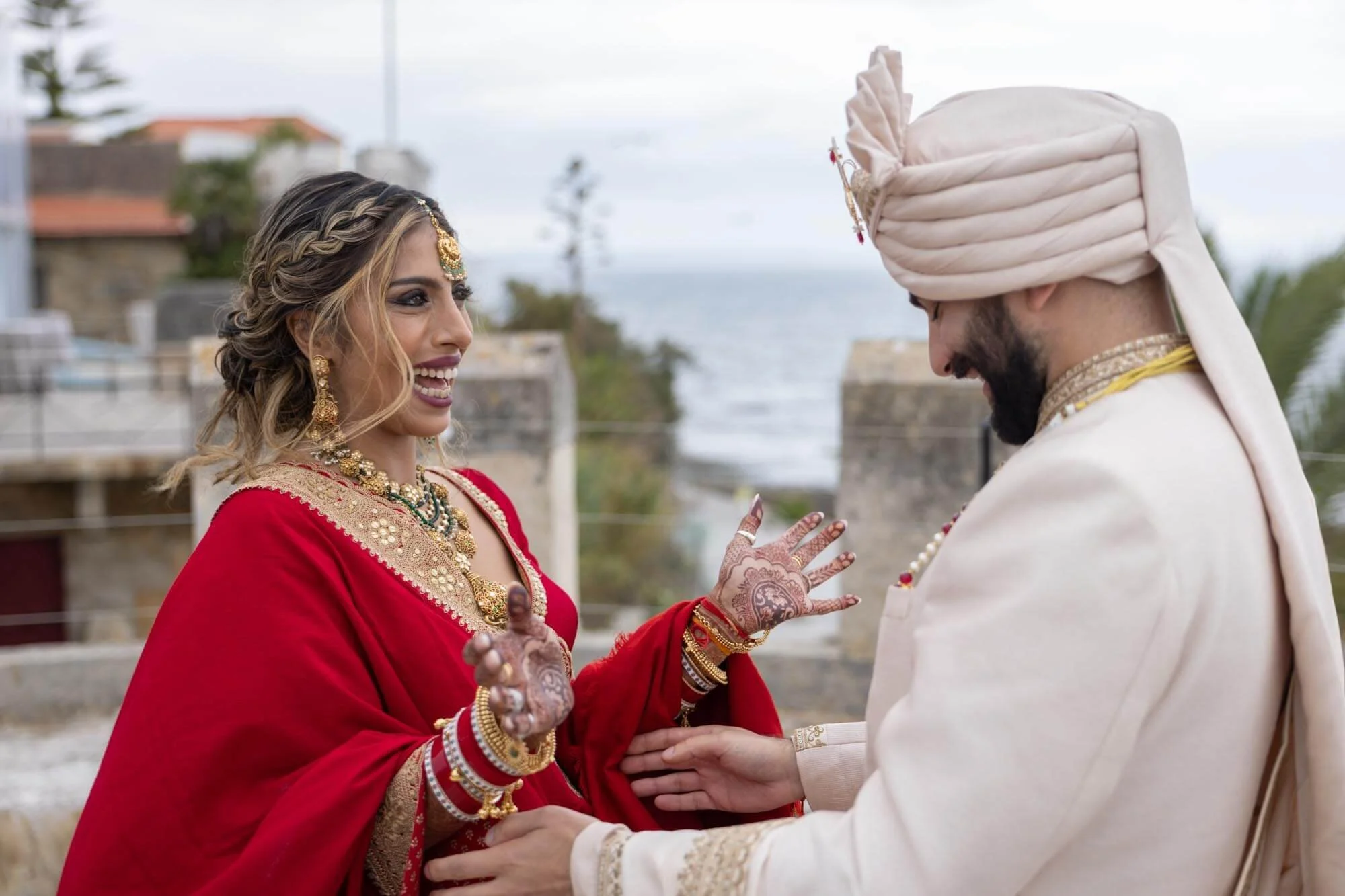 Indian couple in wedding attire sharing a joyful moment outdoors with the ocean and sky in the background.
