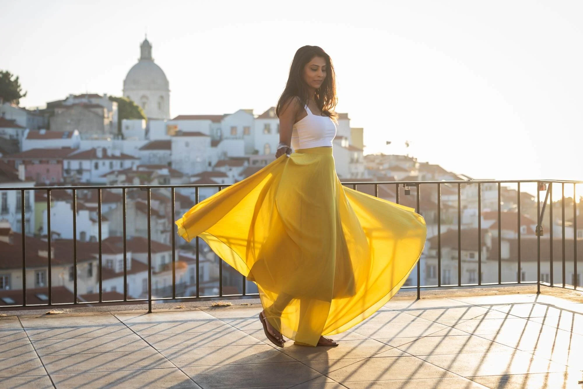 A woman standing on a balcony in a white top and a flowing yellow skirt, with a cityscape and a church dome in the background during sunset.