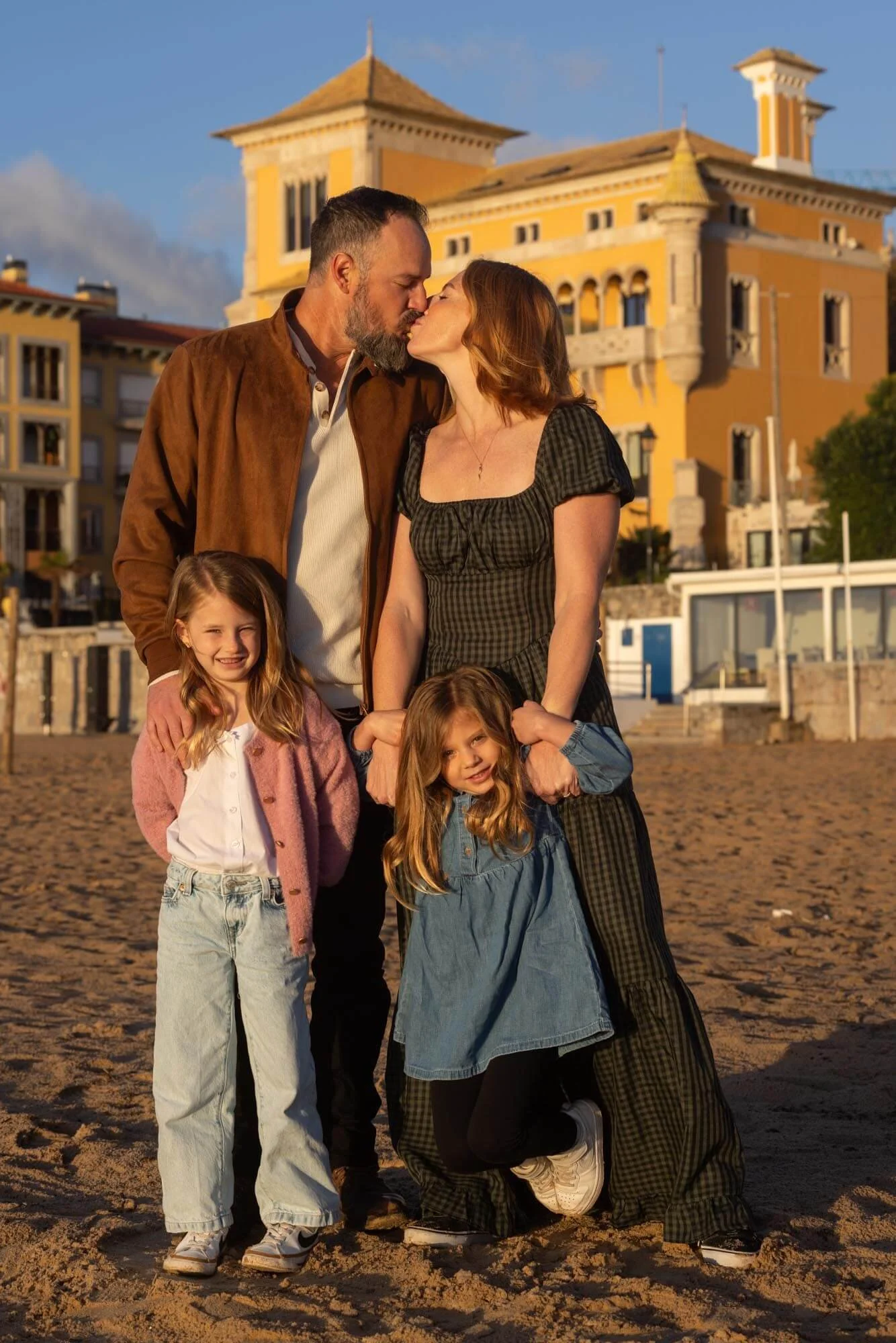 A family of four on a beach during sunset, sharing a kiss, with colorful buildings in the background.