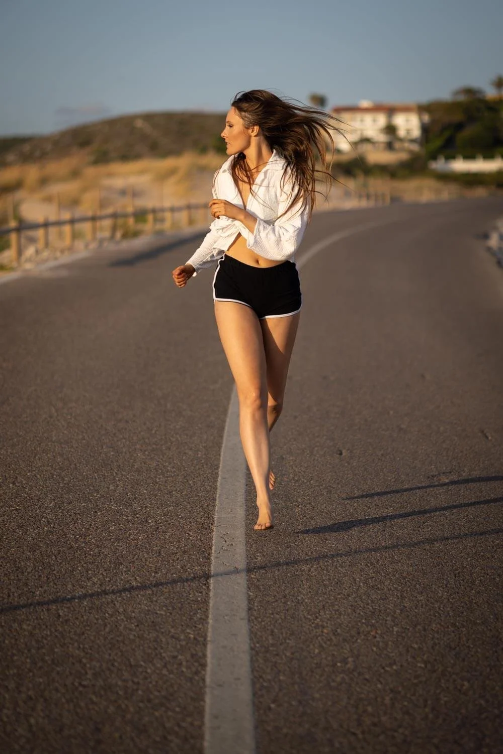 A woman barefoot, running on a deserted road with a white line, wearing a white shirt and black shorts, with her hair flowing in the wind.
