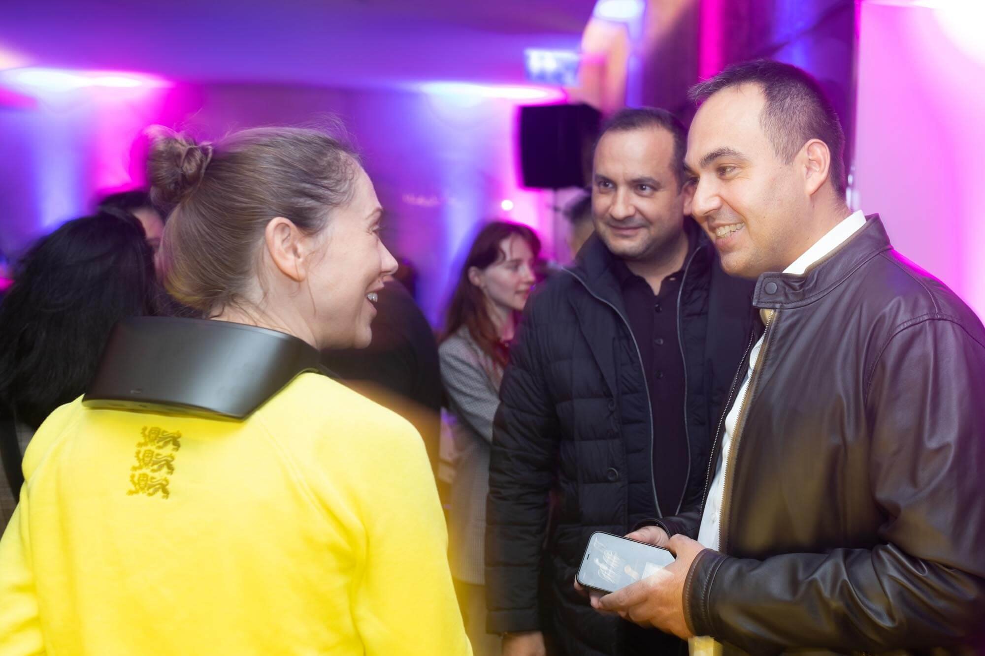 People smiling and talking at an indoor social event with colorful lighting, including a woman in a yellow jacket and a man in a black leather jacket holding a phone.