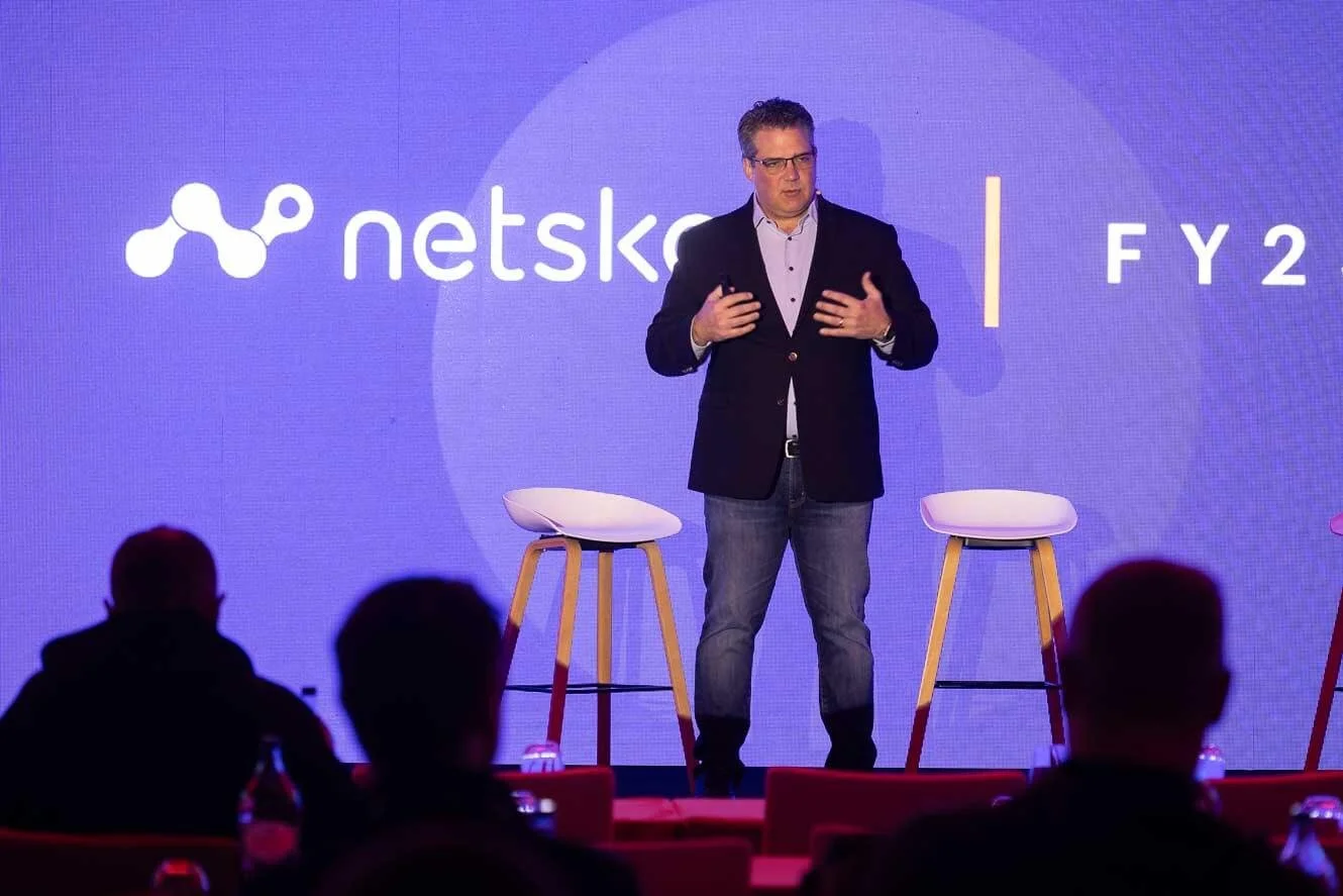 A man in a dark blazer and jeans giving a presentation on stage with a purple background that displays 'netska' and 'FY 2' along with a logo.