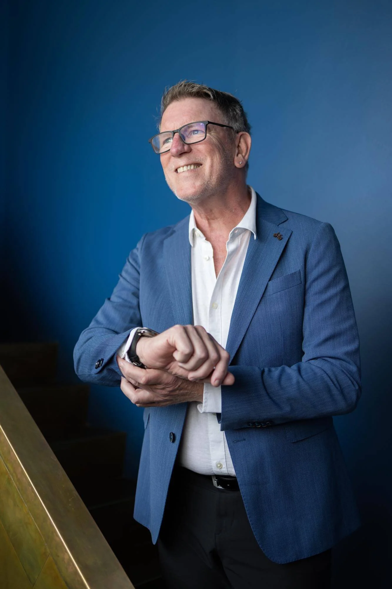 A smiling middle-aged man wearing glasses, a white shirt, and a blue blazer, looking at his watch against a blue background.