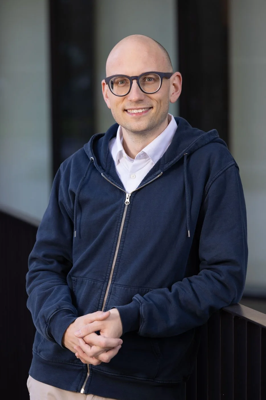 A smiling man with glasses, wearing a white shirt and navy blue hoodie, standing outdoors in front of a building.