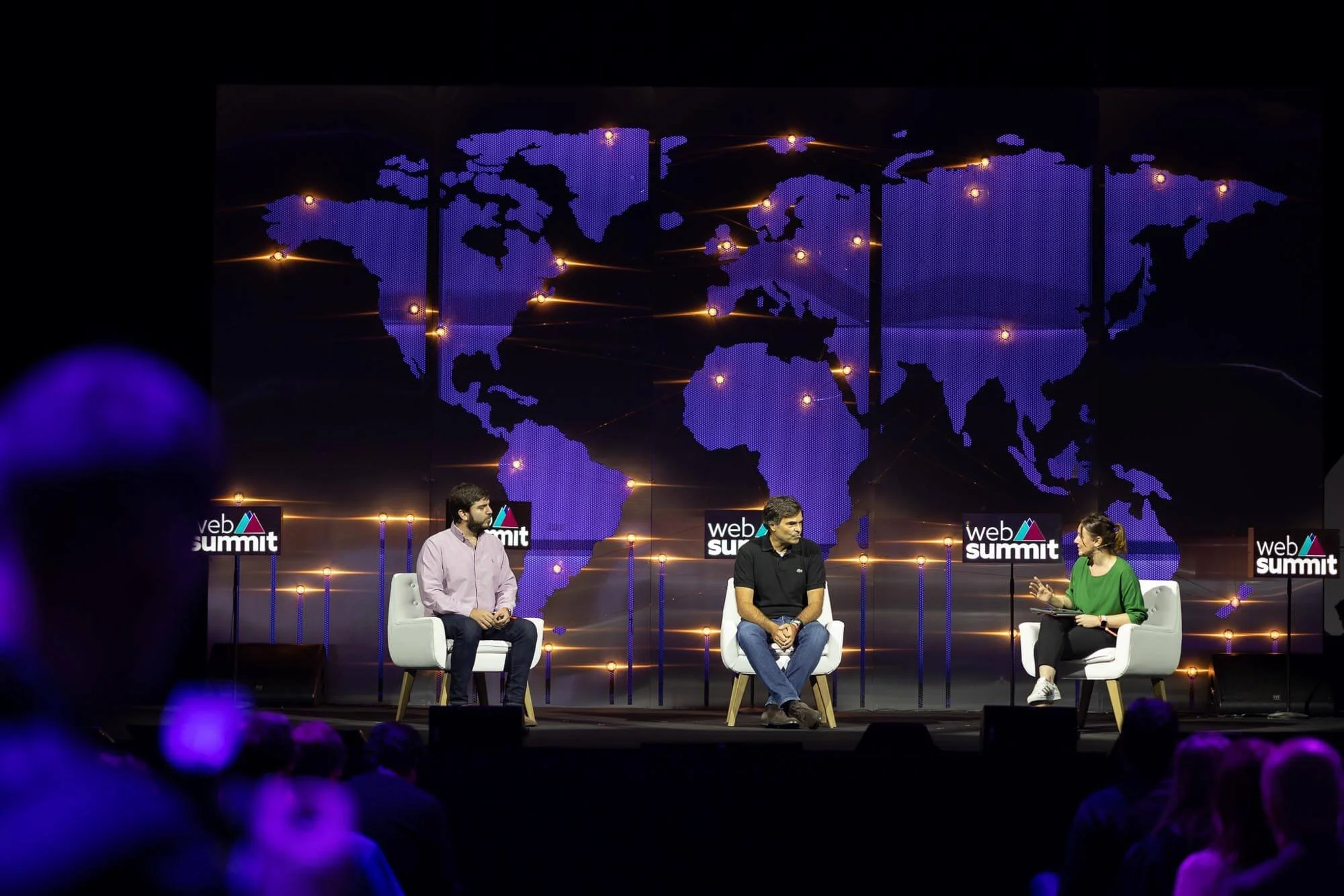 A panel discussion at a web summit with three people on stage, a large world map with illuminated points in the background, and the web summit logo on screens behind them.