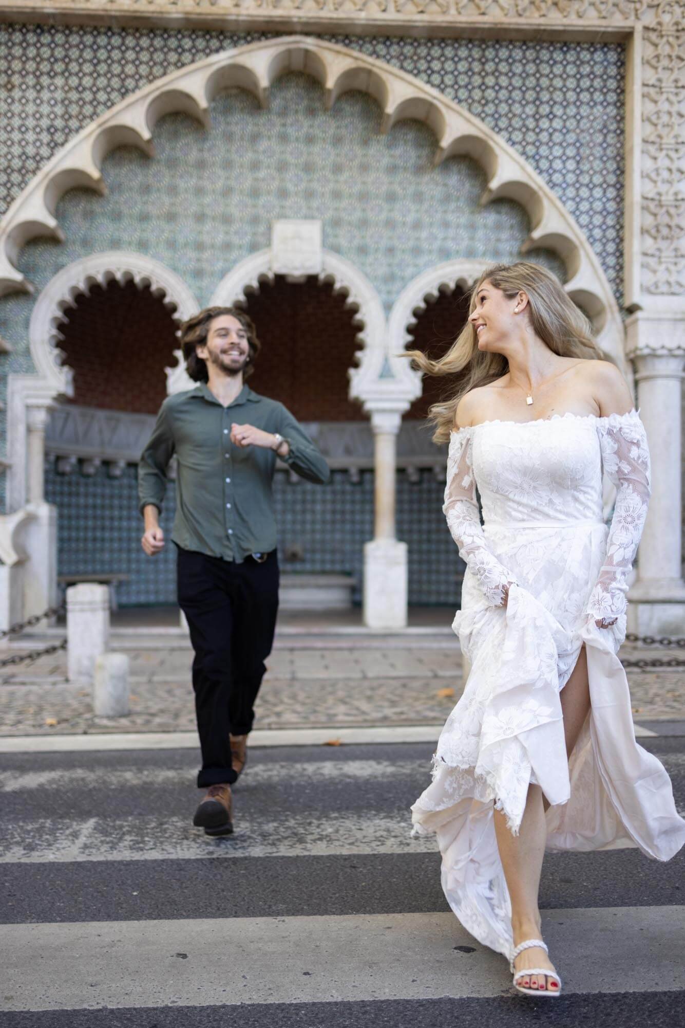 A woman in a white lace off-shoulder dress walking across a street with a man chasing her behind. They are in front of an ornate, patterned building entrance with arches and columns.