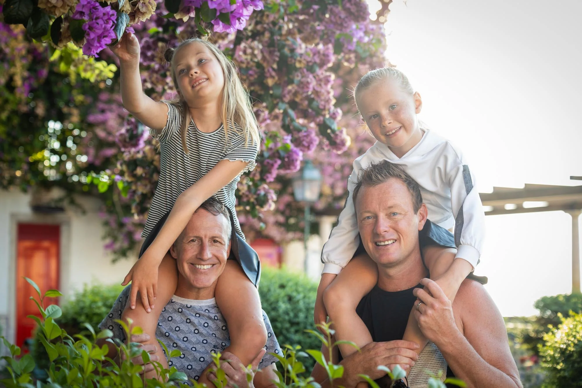 Two men carrying young girls on their shoulders outdoors with pink flowering trees in the background.