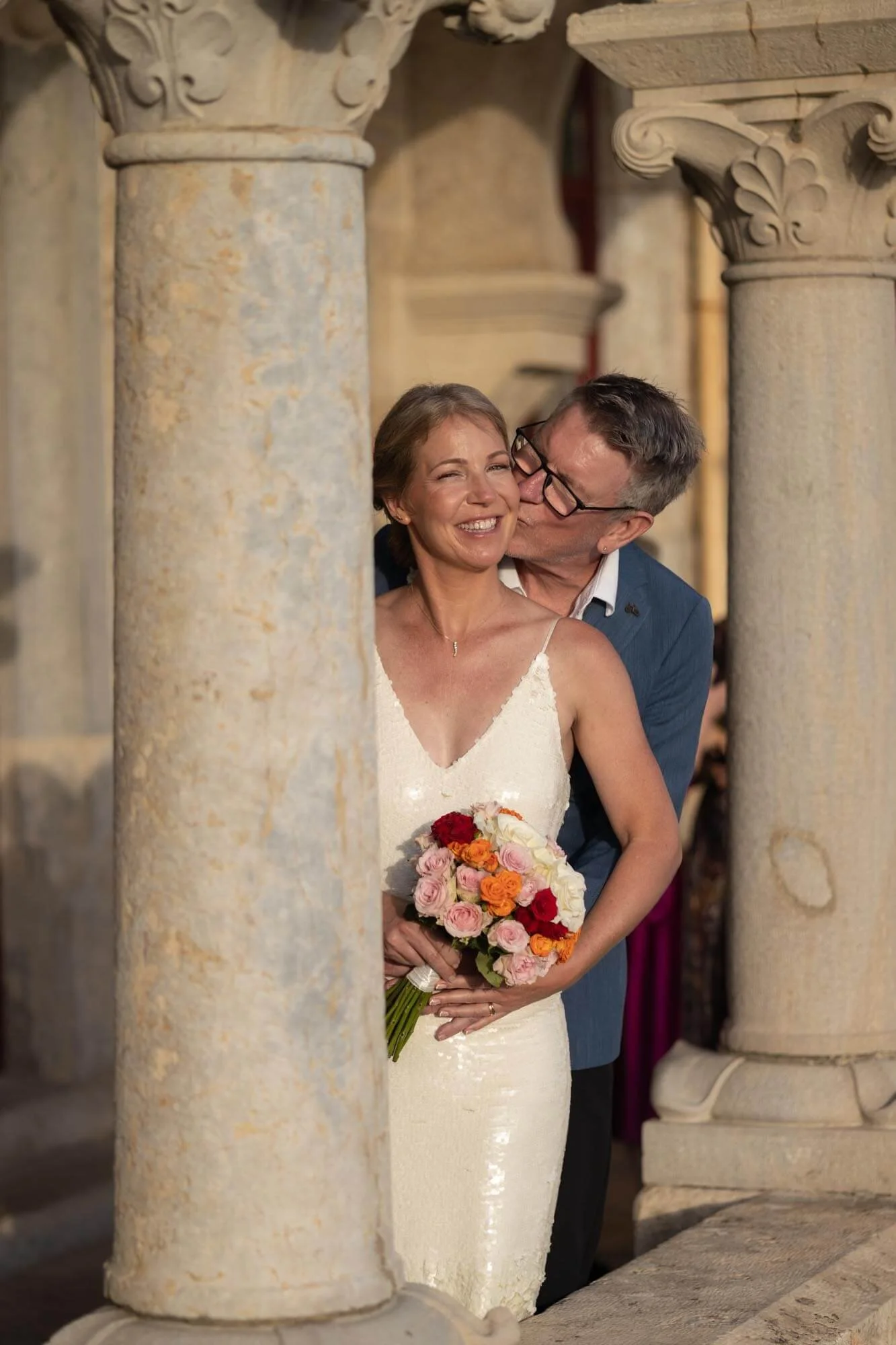 A smiling woman in a white wedding dress holding a colorful bouquet, being kissed on the cheek by a man in glasses and a blue suit during a wedding ceremony.