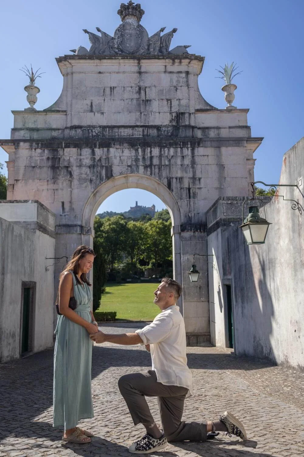A man proposing marriage to a woman in an outdoor historic archway in a park, with a castle visible in the background on a clear, sunny day.