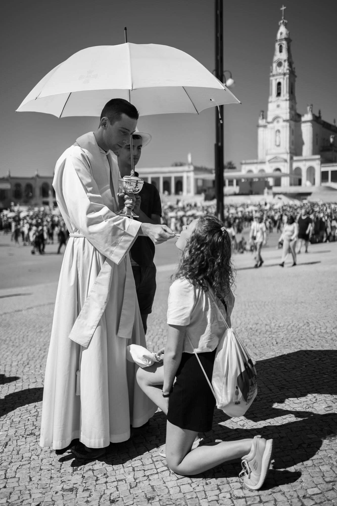 A priest in a white robe unties a woman's shirt collar while she kneels on the ground, with a large crowd in the background.