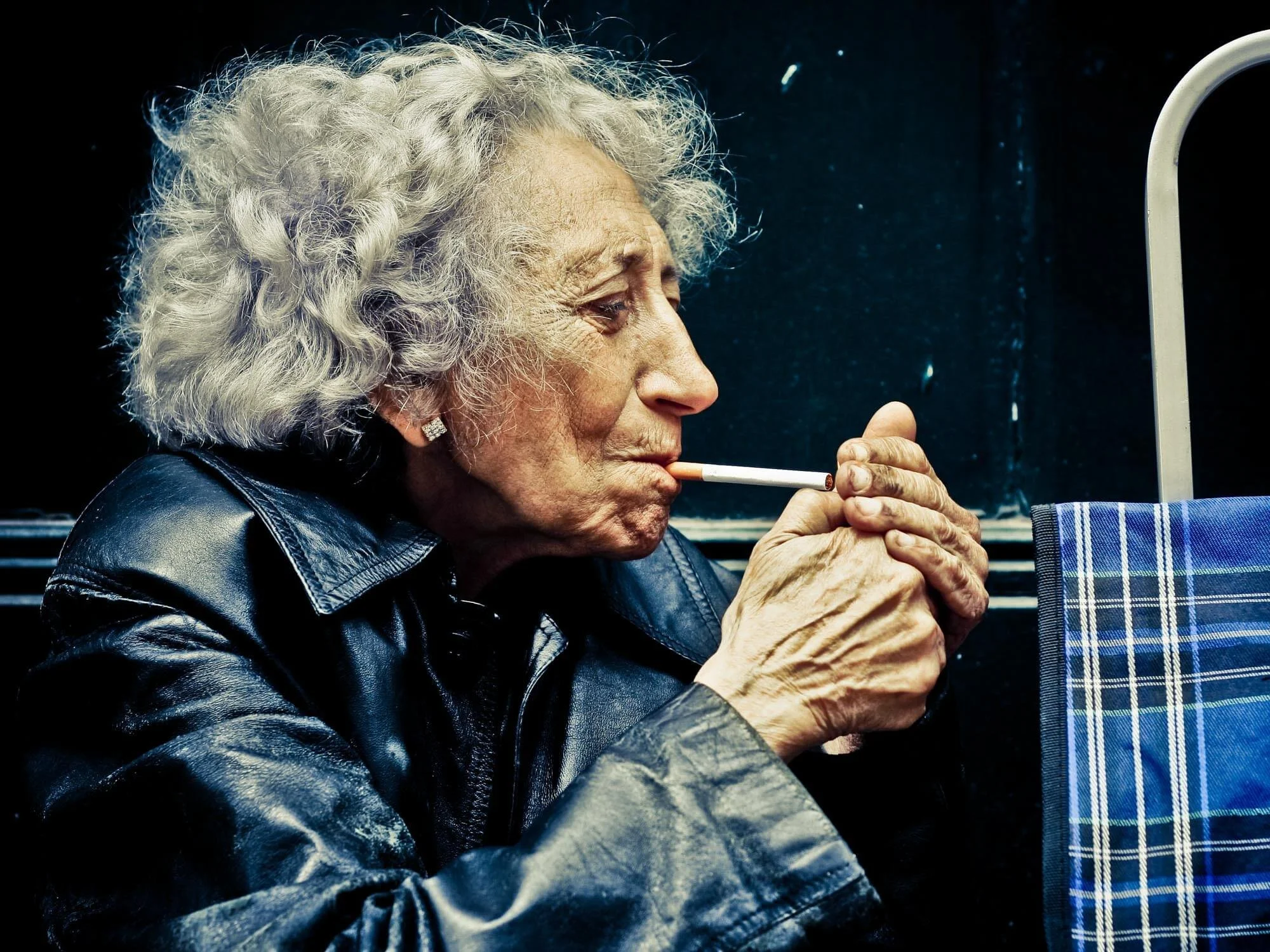 An elderly woman with curly gray hair, wearing a black leather jacket, is smoking a cigarette on a bus or train, with her hands clasped near her mouth.
