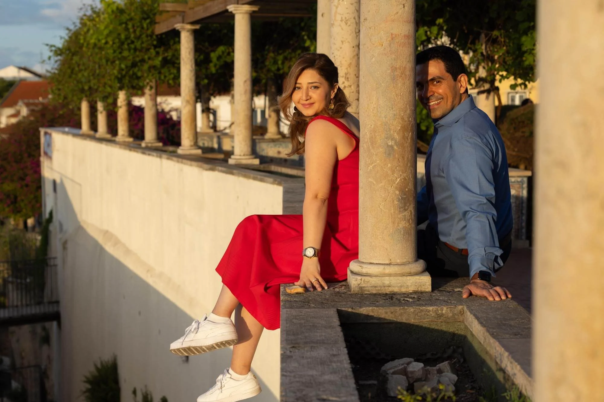 A woman in a red dress and white sneakers sitting on a ledge, with a man in a blue shirt behind a pillar, both smiling outdoors during sunset.