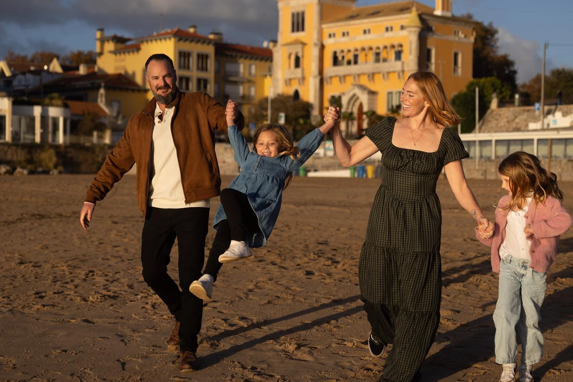 A family of four with two young girls, walking on the beach during sunset, holding hands and smiling, with large yellow and white houses in the background.
