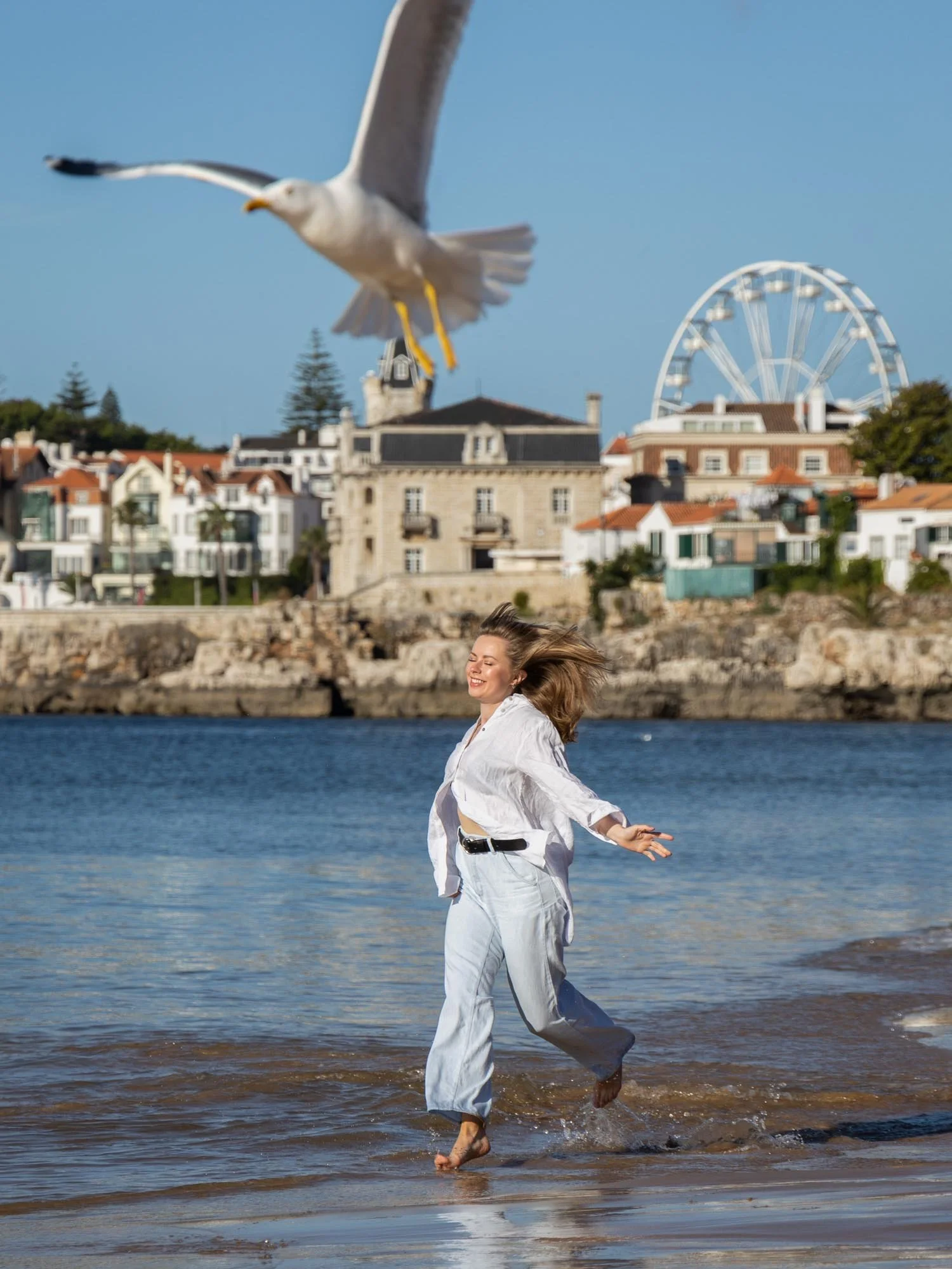 A woman in white pants and shirt joyfully running barefoot along the shoreline with her hair flying, a seagull flying overhead, and a scenic backdrop of buildings, a ferris wheel, and a clear blue sky.