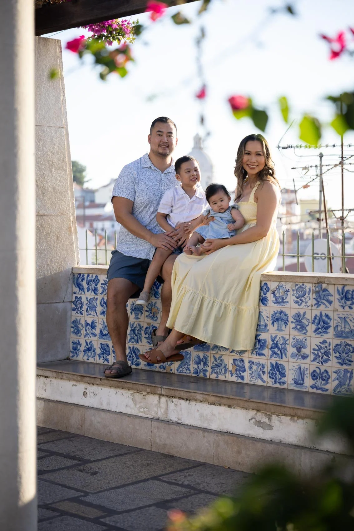 A family of four outdoors posing on a decorative tiled ledge, with pink flowers hanging overhead and a cityscape in the background, during daytime.