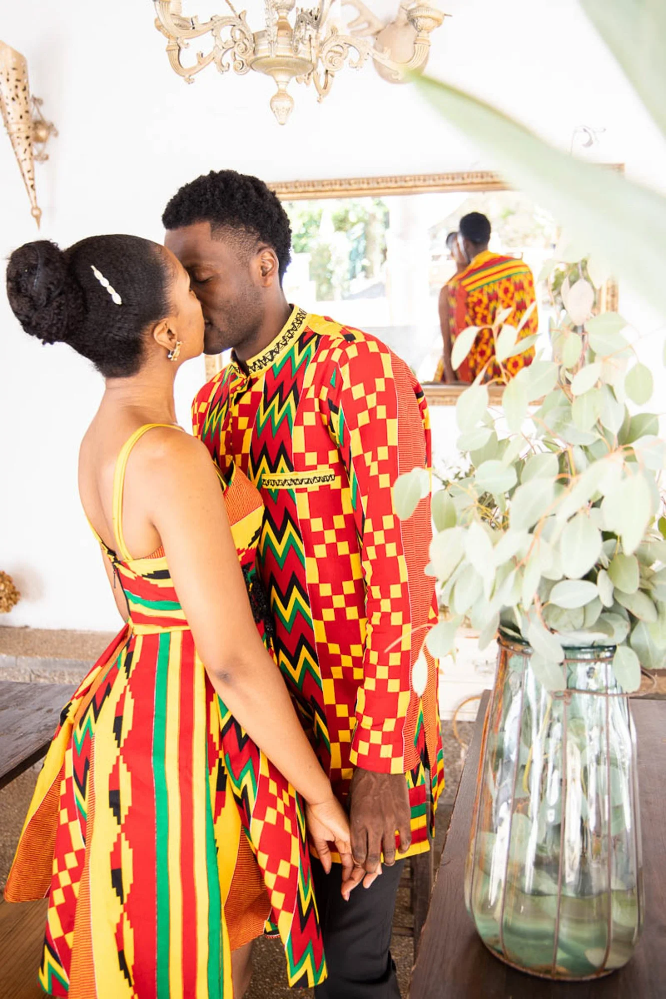 A couple dressed in matching colorful African print outfits sharing a kiss in a well-lit room with a mirror and a vase of greenery.