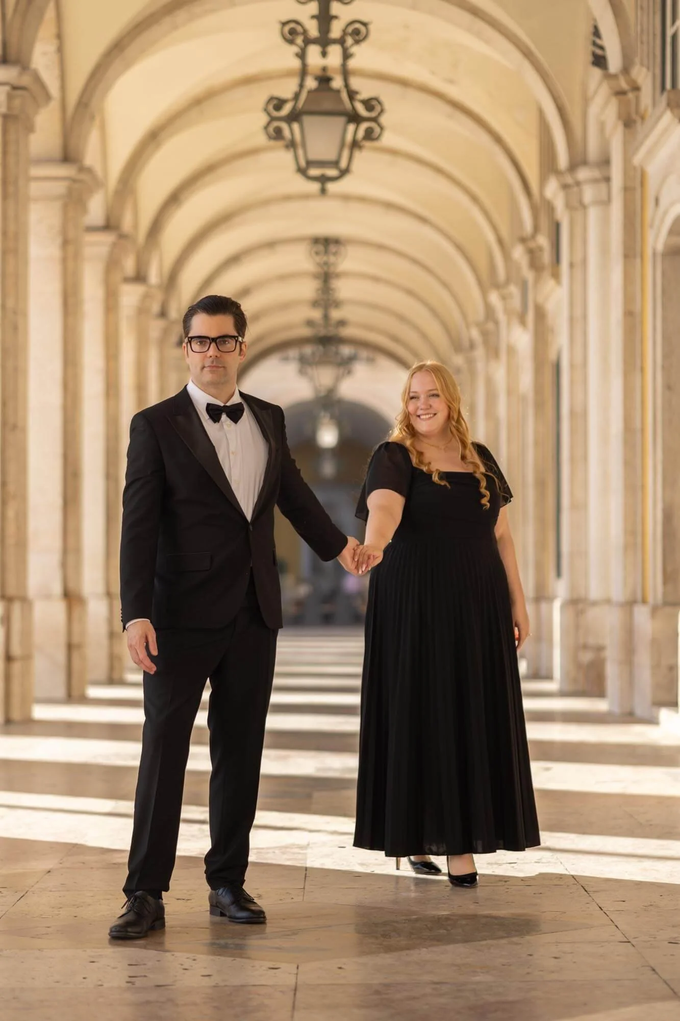 A man in a tuxedo and a woman in a black gown holding hands in an elegant archway, posing for elopement photoshoot in Lisbon downtown.