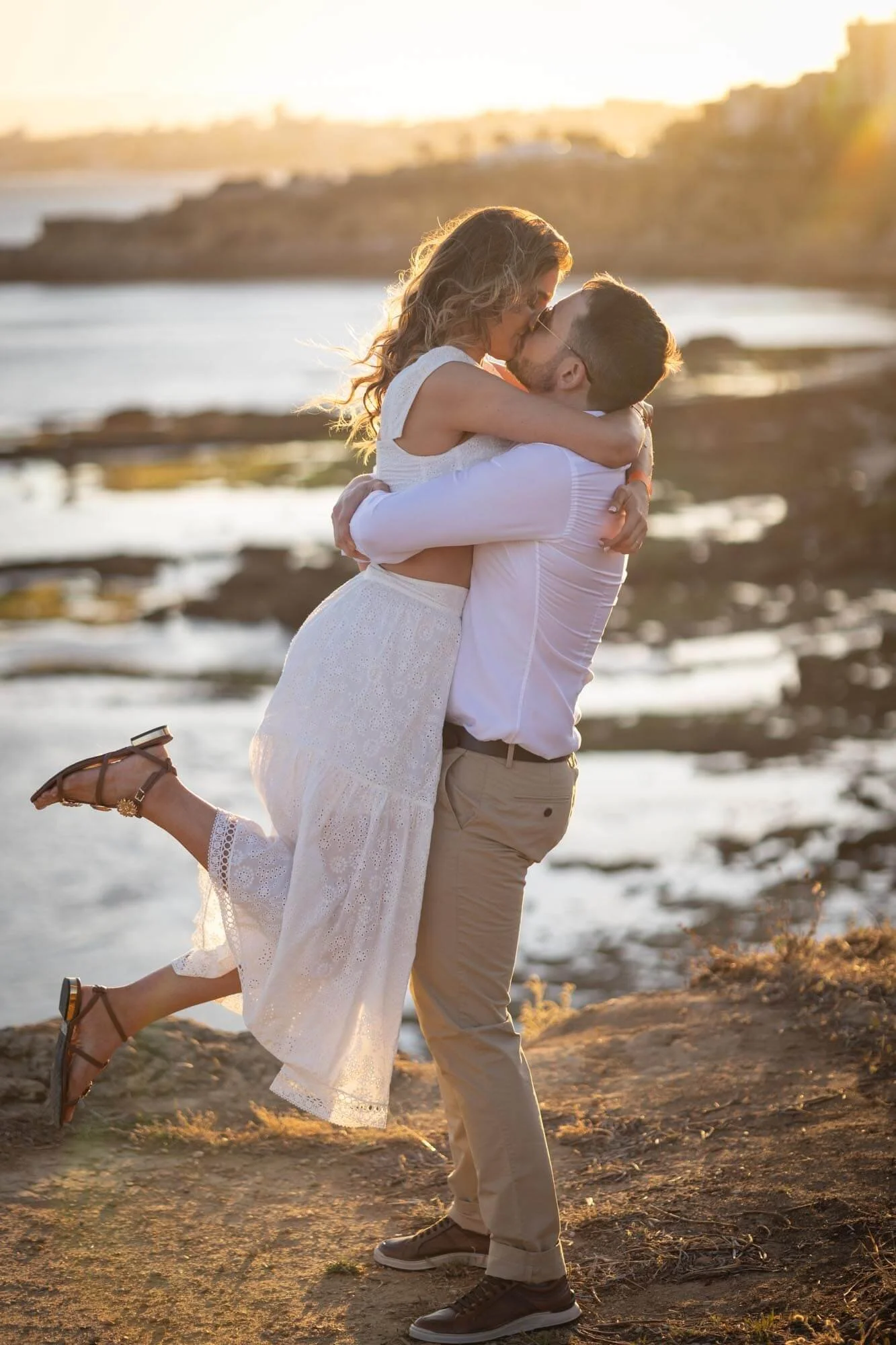 A man lifts a woman in his arms near a rocky shoreline at sunset, sharing a kiss.