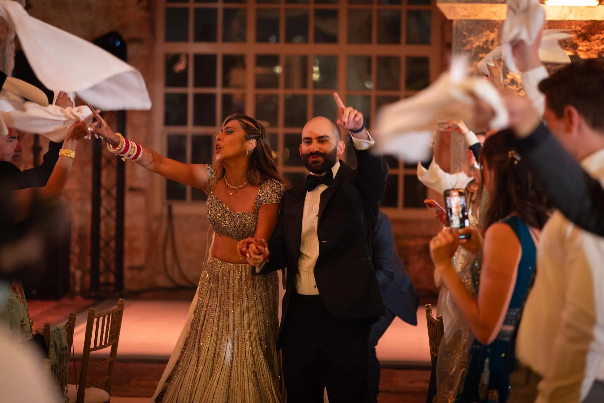 A man in a tuxedo and a woman in a traditional Indian outfit dancing at a celebration or wedding, with guests around them holding white napkins in a warmly lit venue.