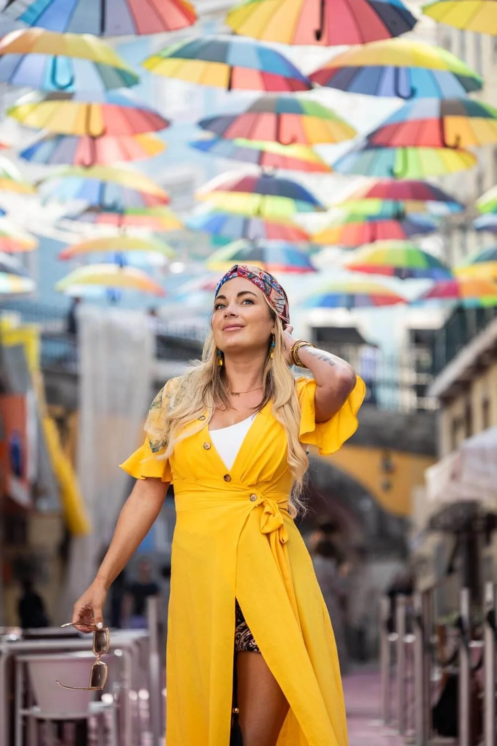 Woman in a yellow dress walking in a colorful city street with hanging rainbow umbrellas overhead.