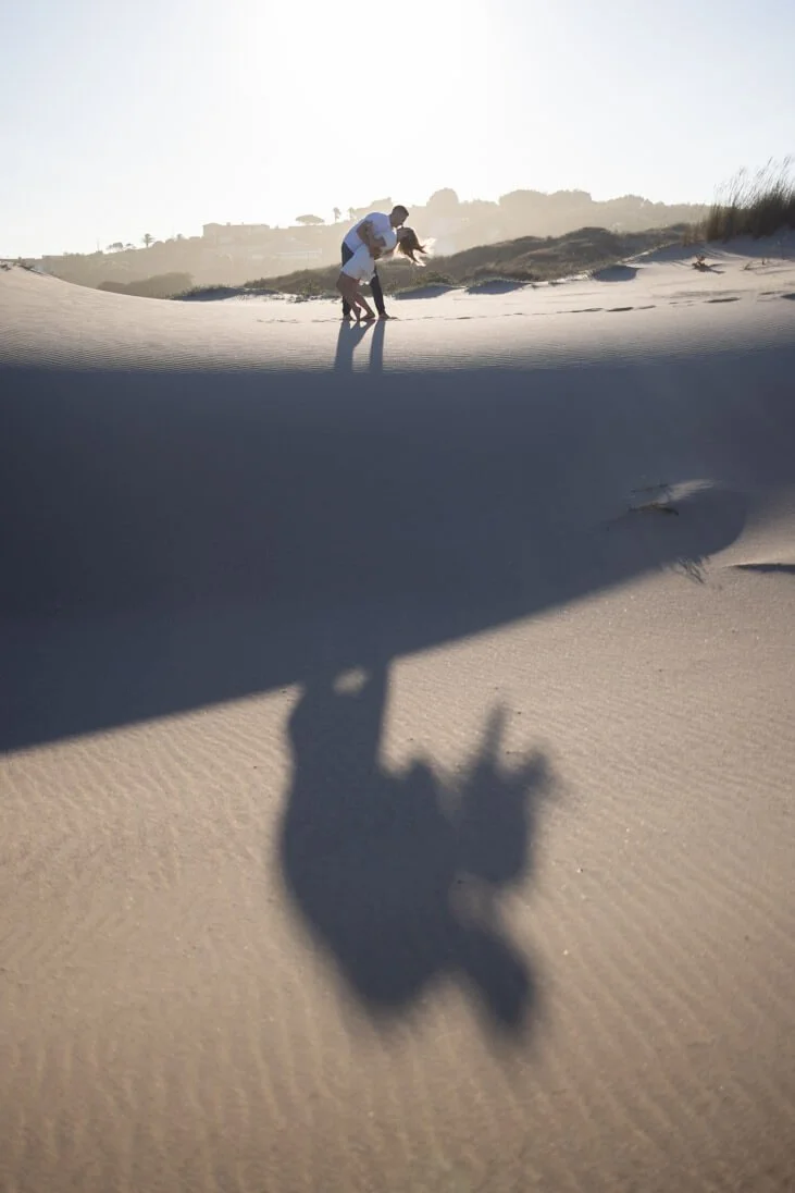 A couple on the beach holds hands and leans over, with the sun casting long shadows on the sand. A crab also casts a shadow nearby.
