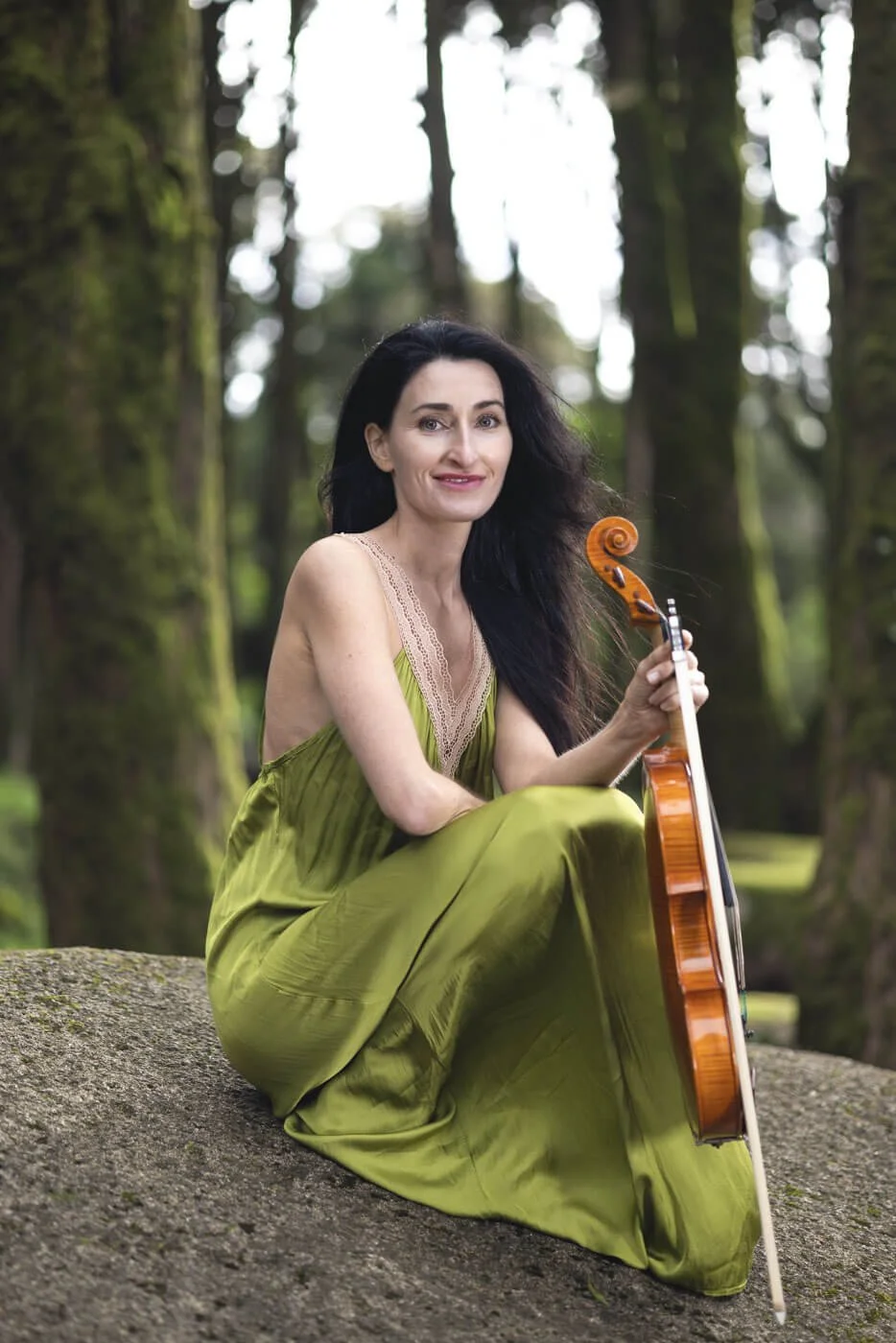 A woman with long black hair sits outdoors on a rock, holding a guitar in a forest with tall trees.
