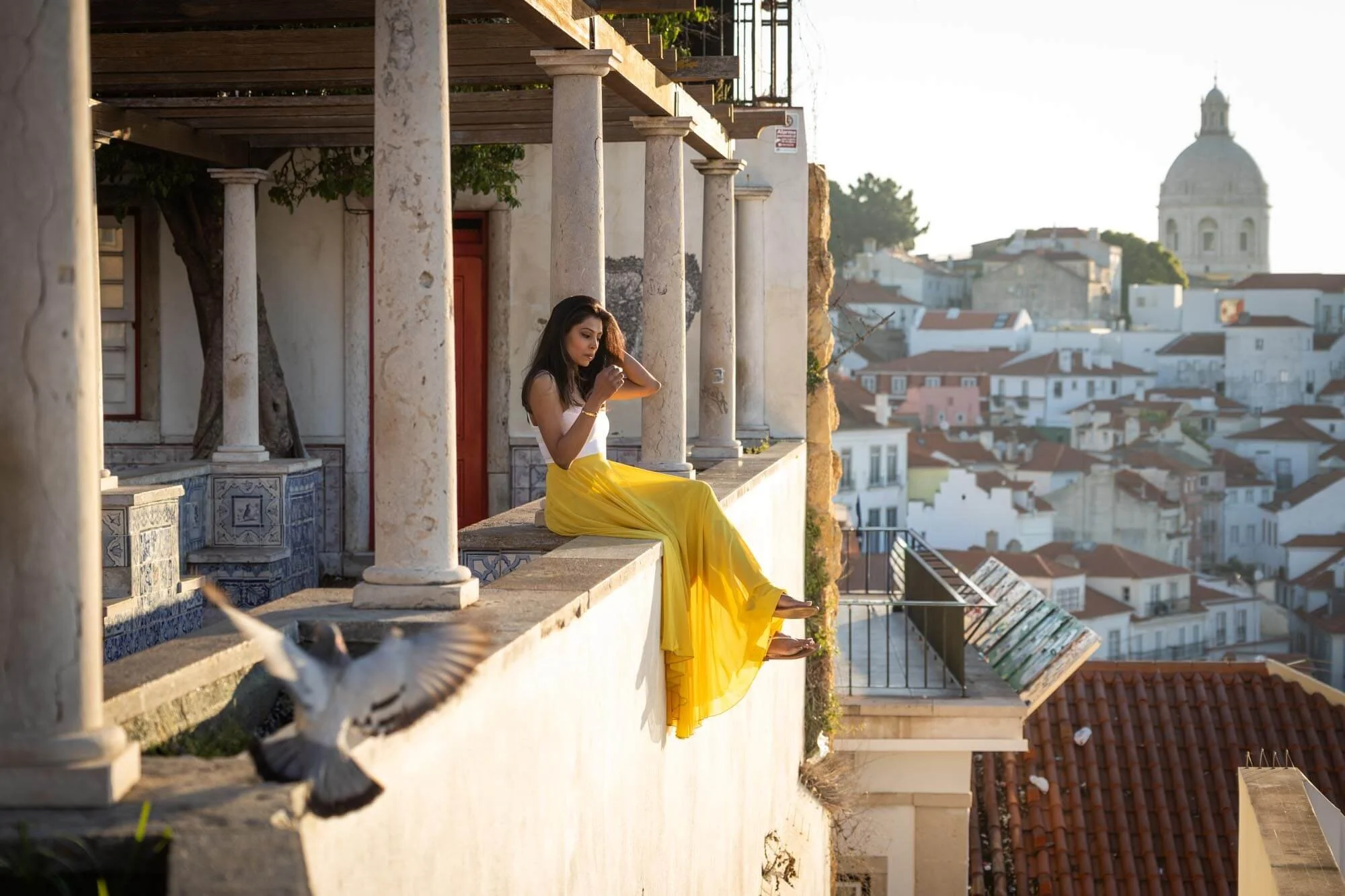 A woman in a white top and yellow skirt sitting on a balcony, reading a letter, with a cityscape featuring a domed building in the background during sunset.