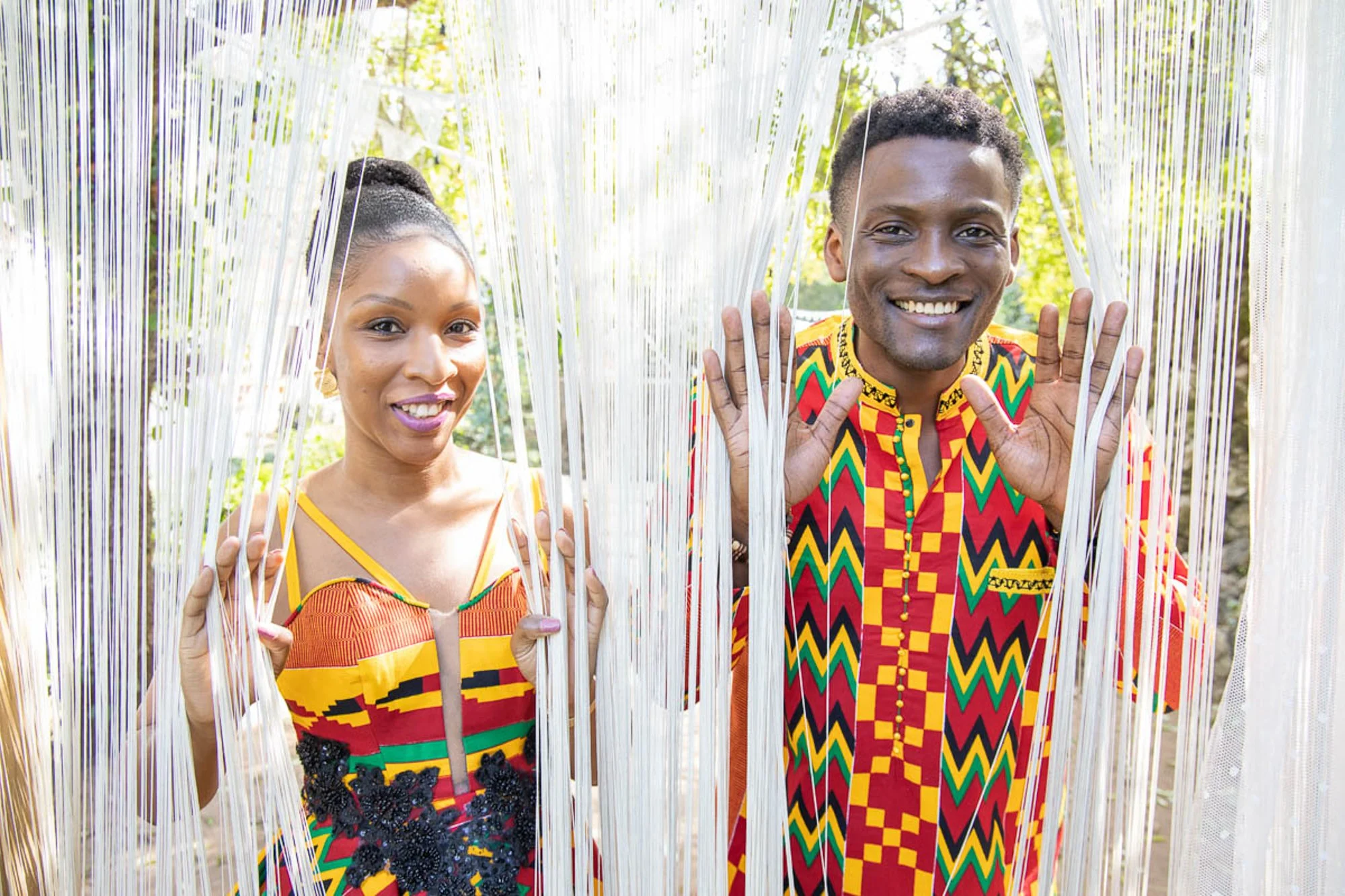 Two people smiling and standing behind white string curtains, wearing colorful African-patterned clothing outdoors.