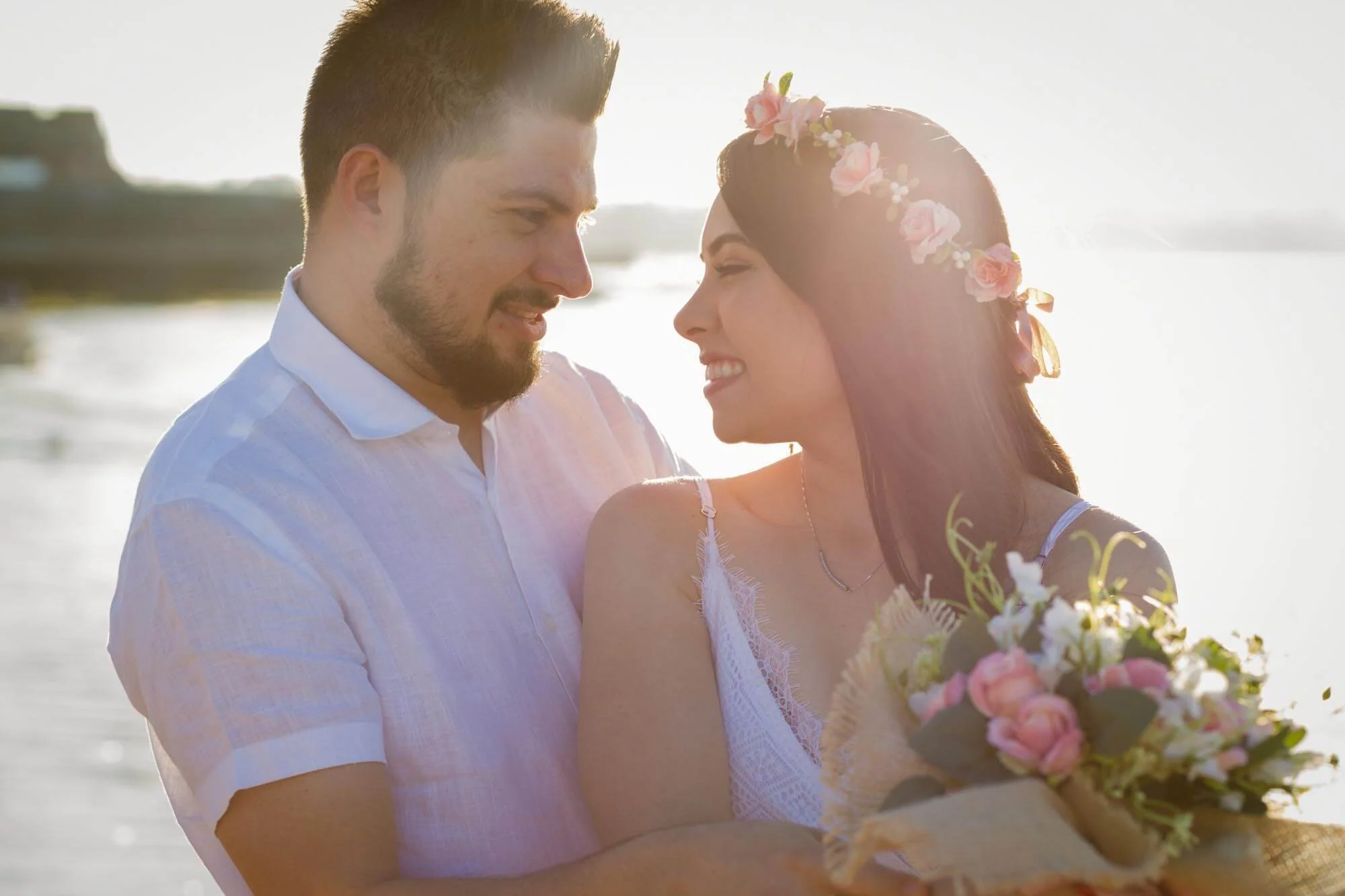 A smiling couple, a man and a woman, on a beach during sunset, with the woman holding a bouquet of flowers and both wearing light-colored clothing.