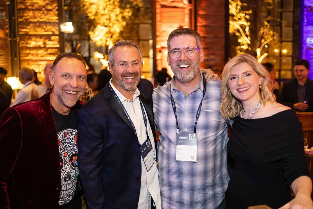 Four people smiling and posing for a photo at a social event or party. They are dressed in smart casual attire, with name tags around their necks. The background shows warm lighting, a brick wall, and other attendees.
