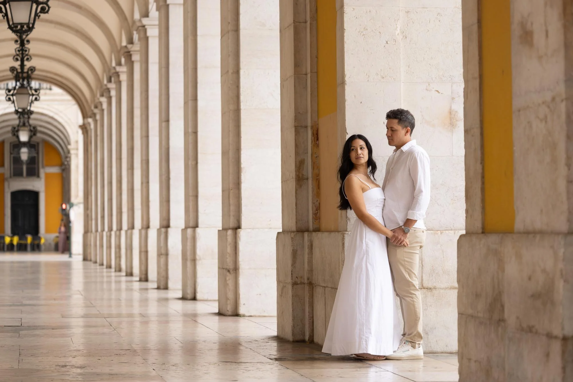 A couple in white dresses standing close together under a colonnade with arches, facing each other.