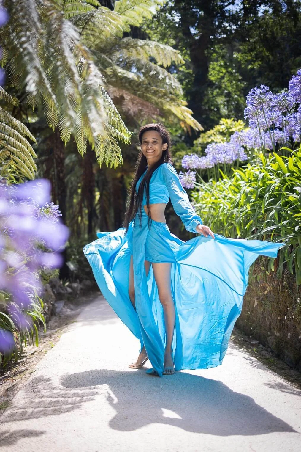 A woman in a flowing blue dress standing on a garden path with lush green foliage and vibrant purple flowers around her, smiling while holding her dress out to the side.