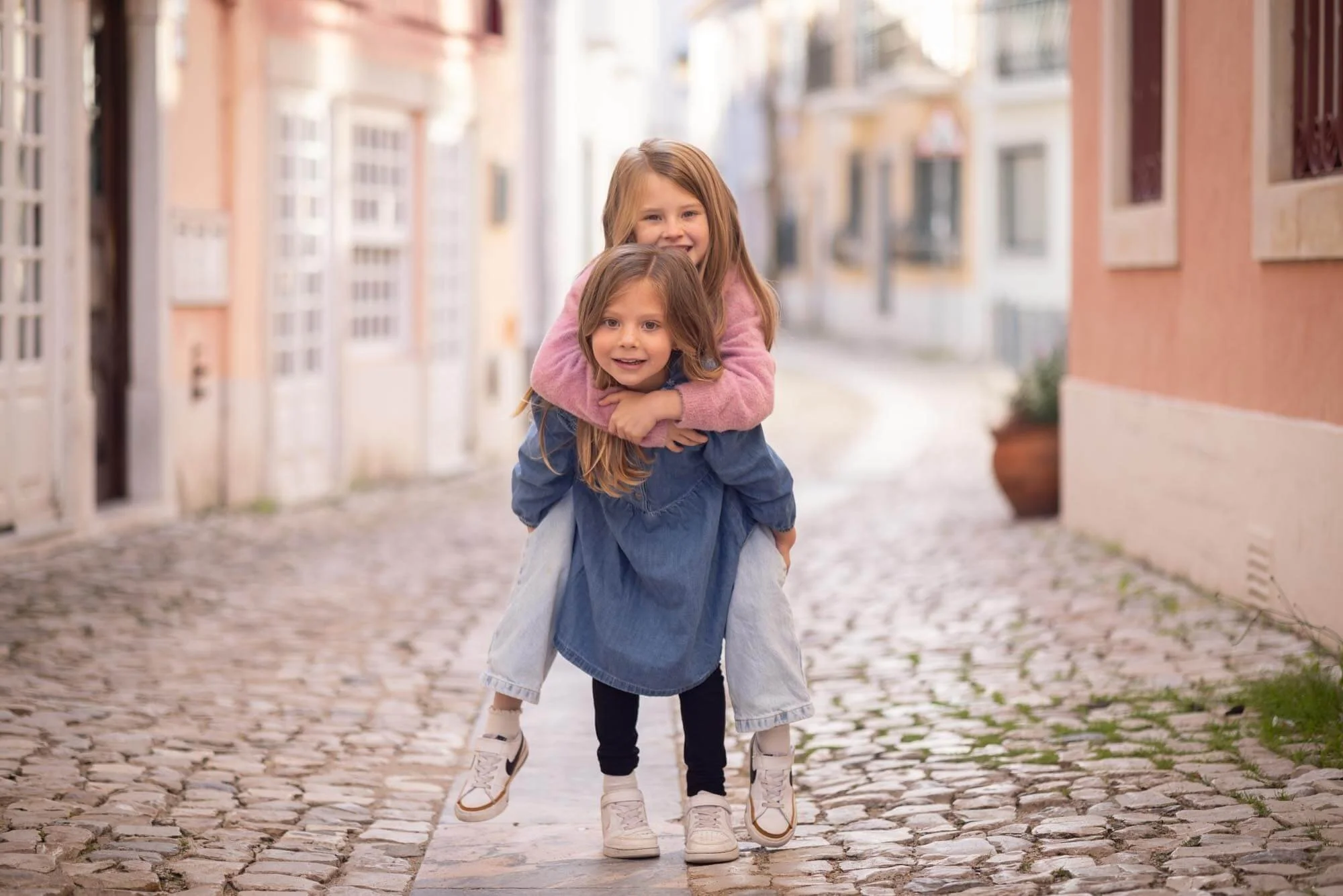 Two young girls are having fun on a cobblestone street, with one girl giving the other a piggyback ride. Both are smiling and enjoying the moment, wearing casual clothing.
