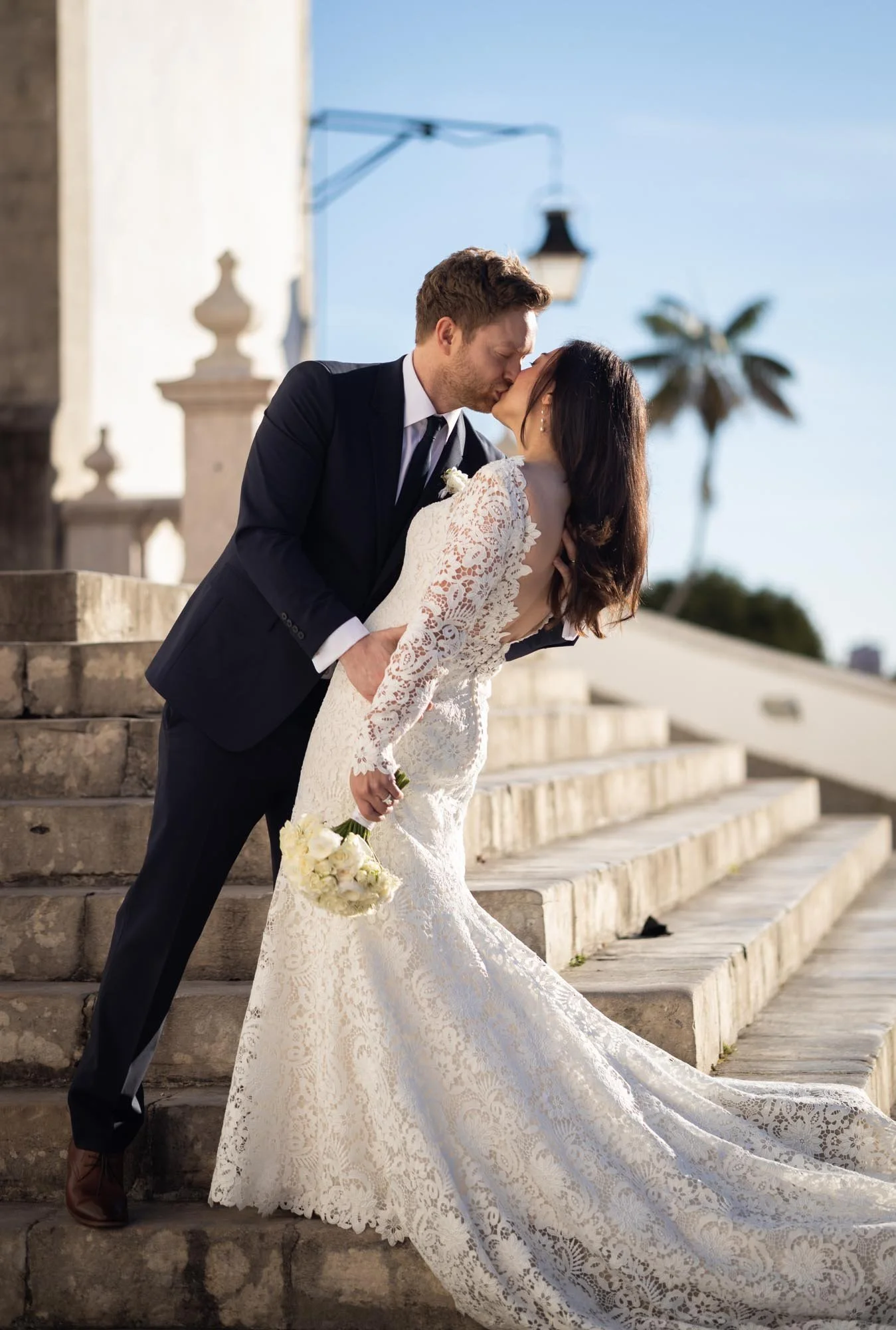 A bride and groom sharing a kiss on a staircase outdoors during their wedding, with the bride holding a bouquet of white flowers, and the bride in a lace wedding dress and the groom in a dark suit.