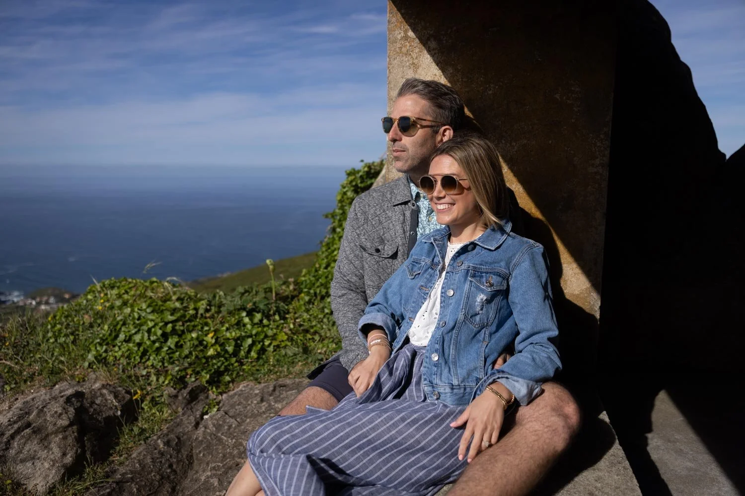 A smiling woman and man sitting outdoors on rocks, with ocean and blue sky in the background, both wearing sunglasses.