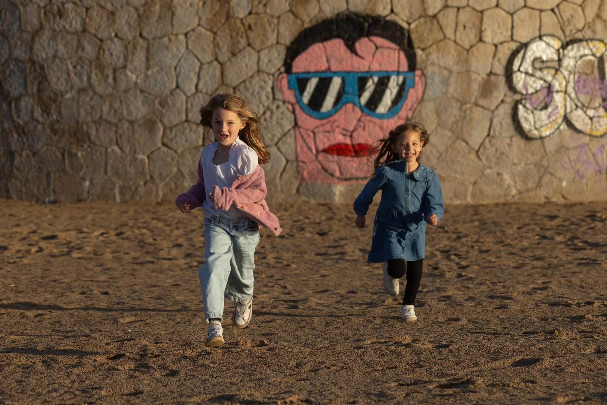 Two young girls running on a sandy surface in front of a stone wall graffiti featuring a face with sunglasses and red lips.